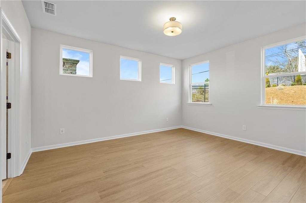 Spacious empty bedroom with hardwood floors, light gray walls, and large windows overlooking greenery in The Edison B home, Loganville, GA