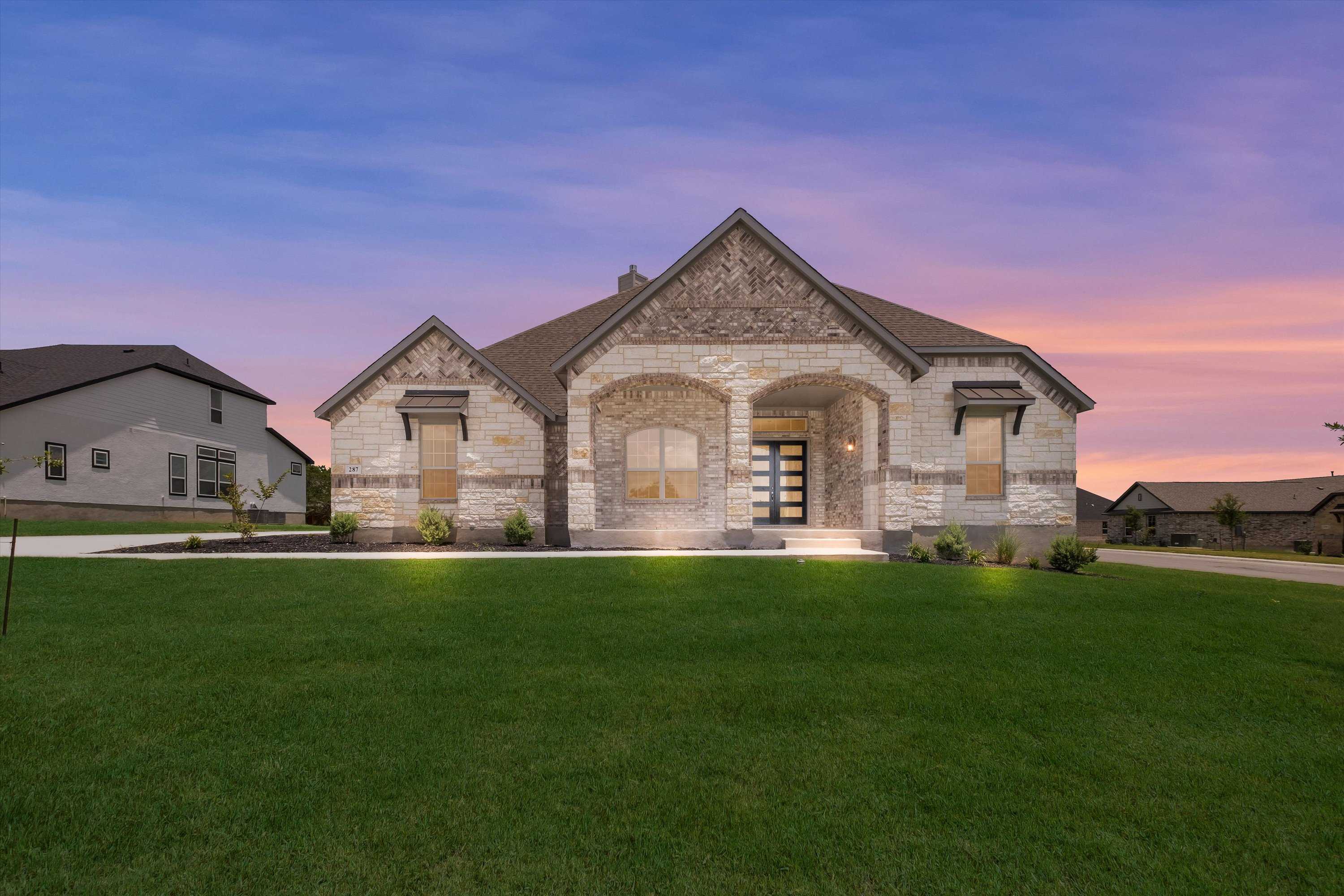 Elegant stone-front single-story home with arched windows, 3-car garage, and landscaped lawn at sunset in Potranco Oaks, Castroville, Texas