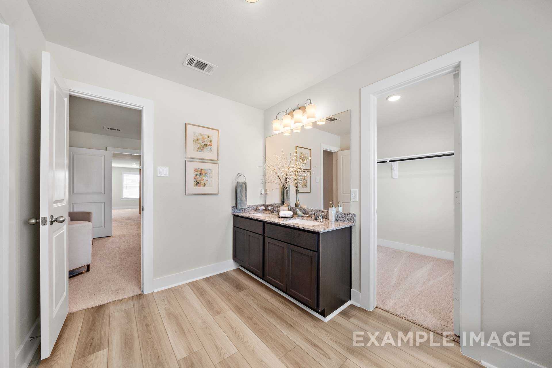 Elegant master bathroom in The Lily A with dark wood double vanity, lighted mirror, and walk-in closet access
