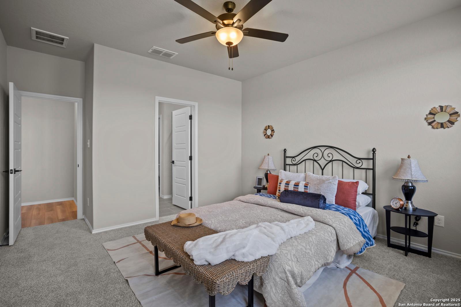 Inviting bedroom with metal headboard bed, plush bedding, ceiling fan, and open door to ensuite in Davidson Homes The Asheville H, Converse, TX