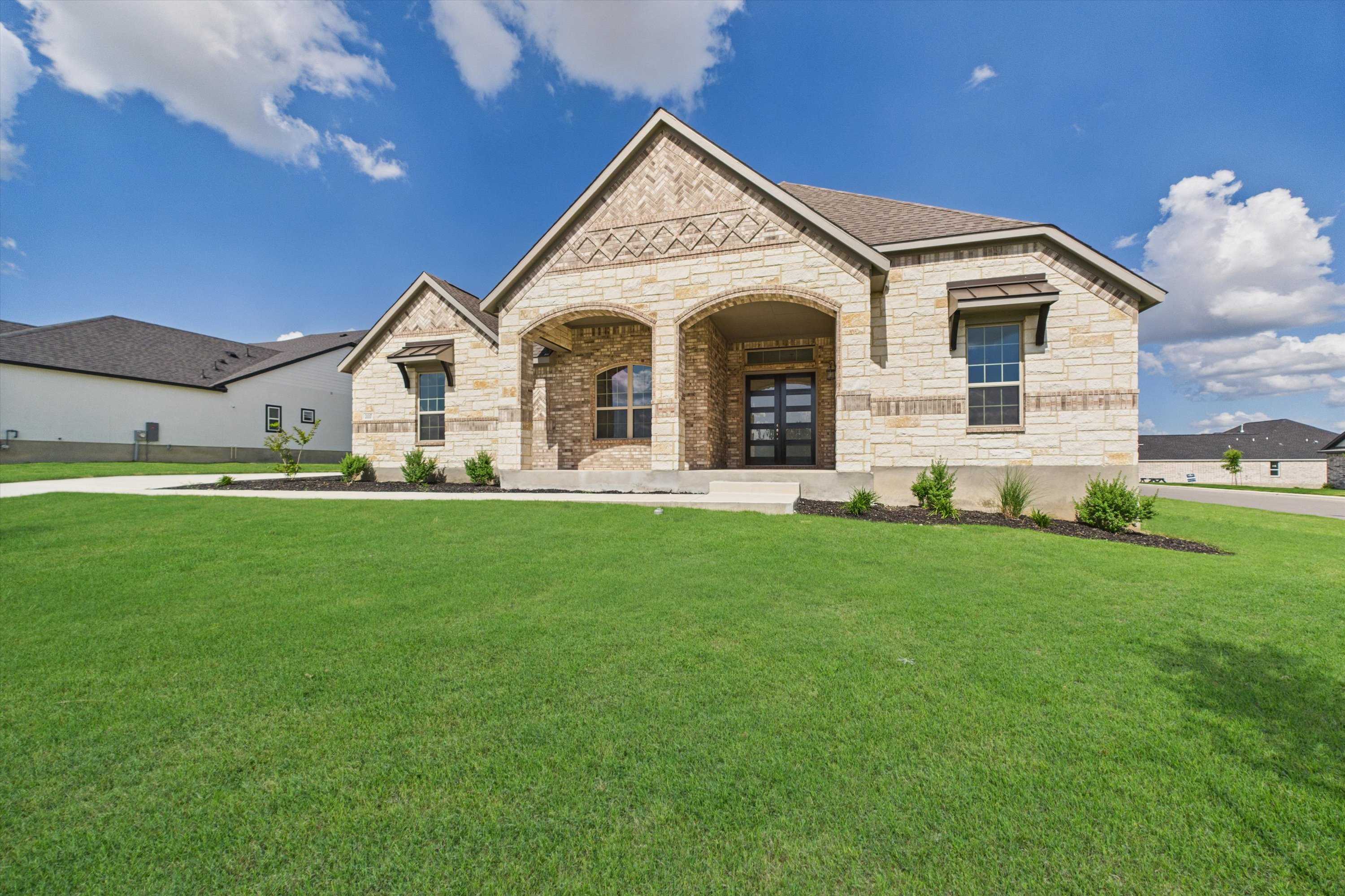 Modern single-story stone home with arched entry, lush green lawn, and blue sky in Potranco Oaks, Castroville, Texas