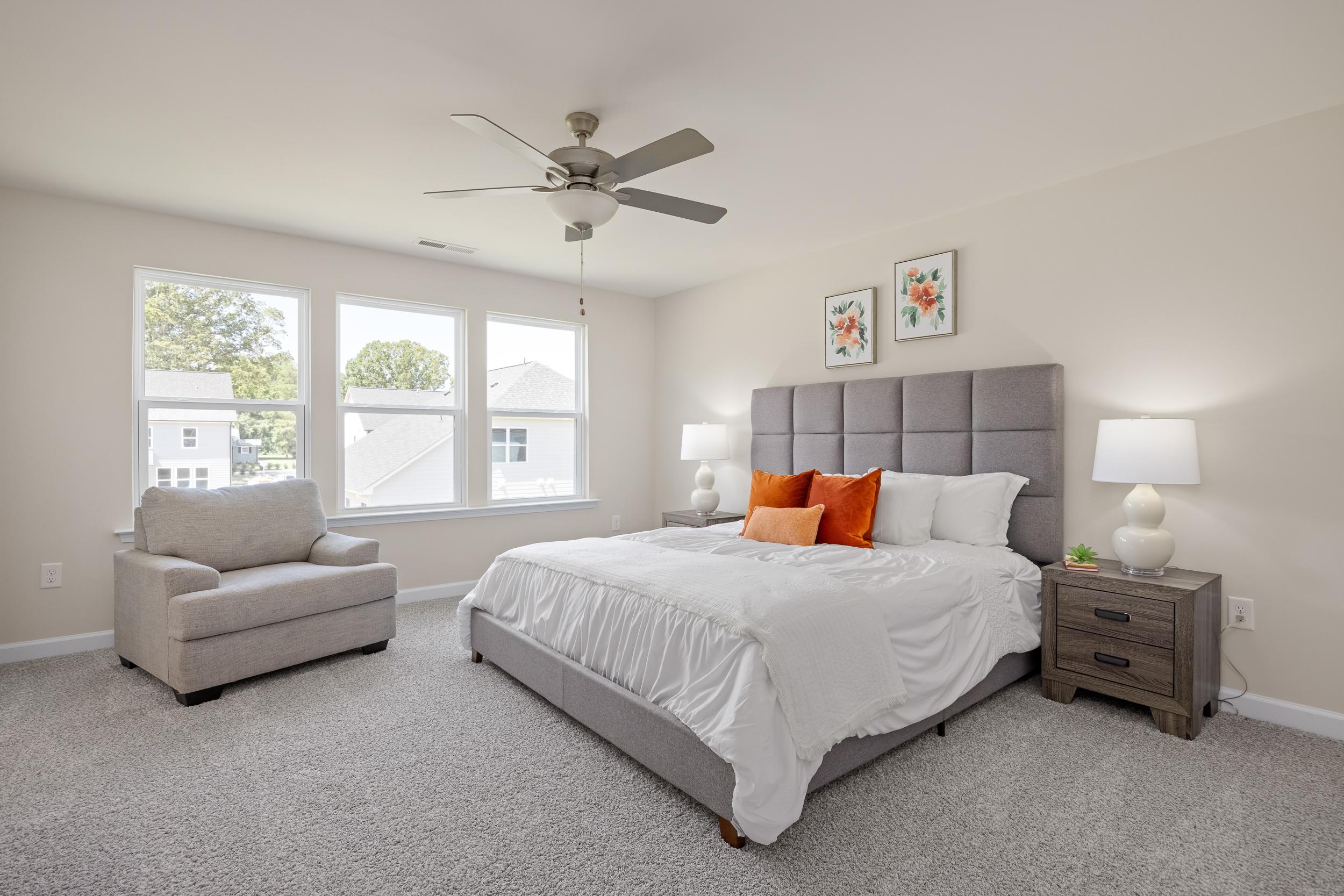 Spacious primary bedroom in Montrose Aberdeen NC with gray tufted headboard orange accent pillows ceiling fan and large windows