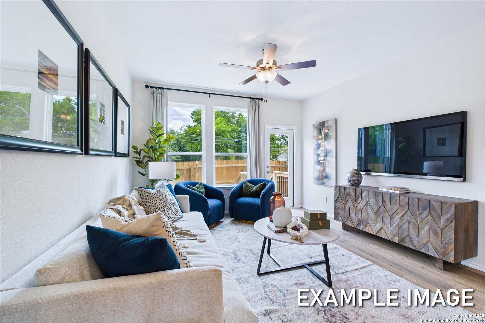 Cozy modern living room with beige sofa, blue velvet chairs, wooden media console, and large garden-view windows in Davidson Homes The Gillian C, San Antonio