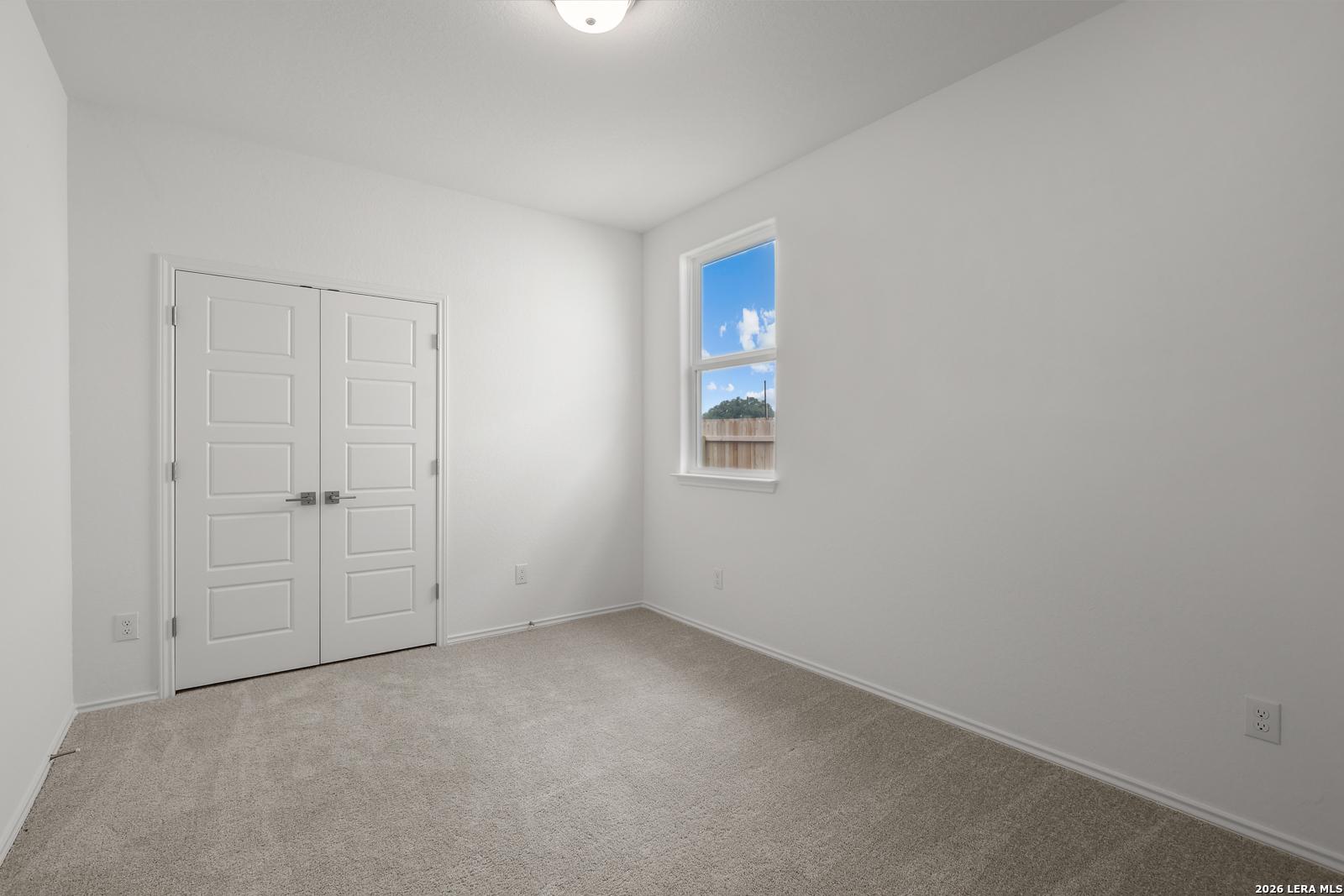 Bright secondary bedroom with white walls, beige carpet, double closet doors, and window view of backyard in The Glenwood E, Elmendorf, Texas