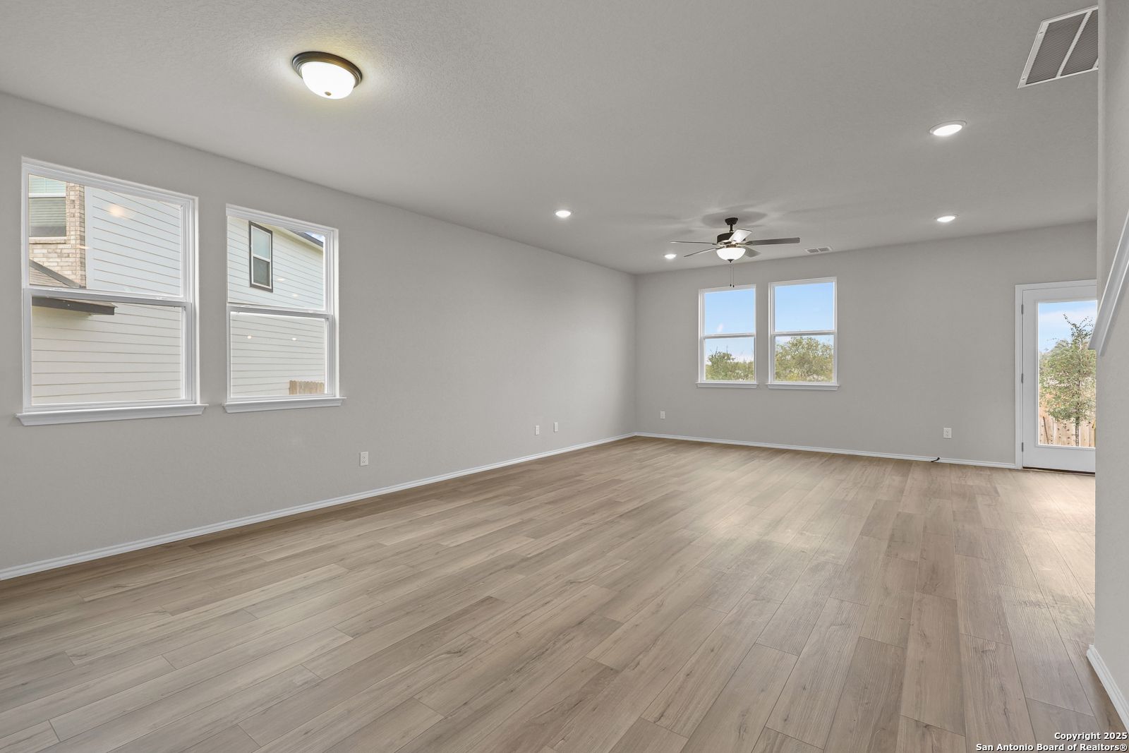 Spacious living room with light gray walls, hardwood floors, large windows, ceiling fan, and sliding door in Davidson Homes The Murray J, San Antonio