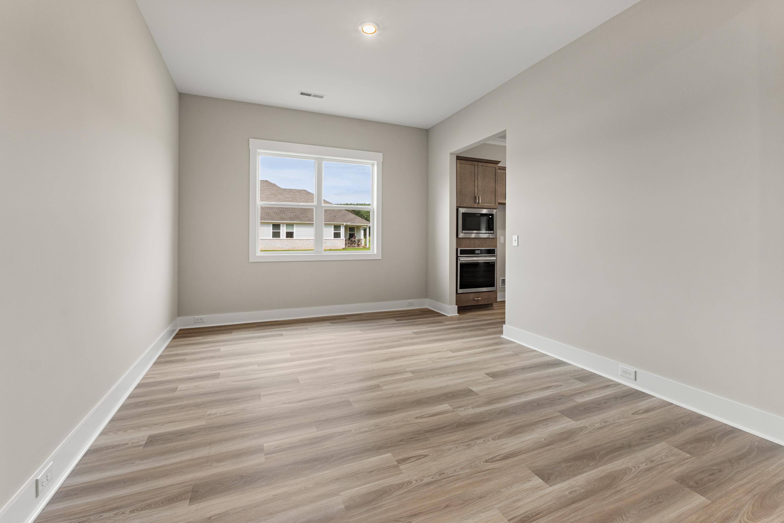 Bright bedroom in The Oxford C with neutral walls, large window to neighborhood view, adjacent kitchen cabinets, hardwood floors