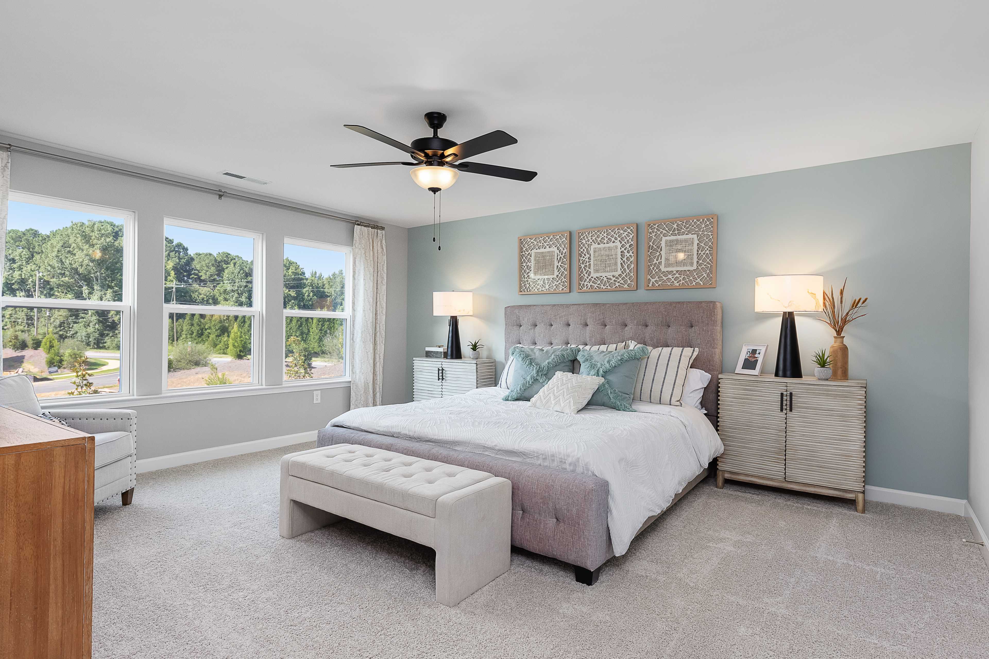 Spacious primary bedroom at Woodland Crossing in Zebulon NC with tufted headboard, plush bedding, ceiling fan, and wooded window views