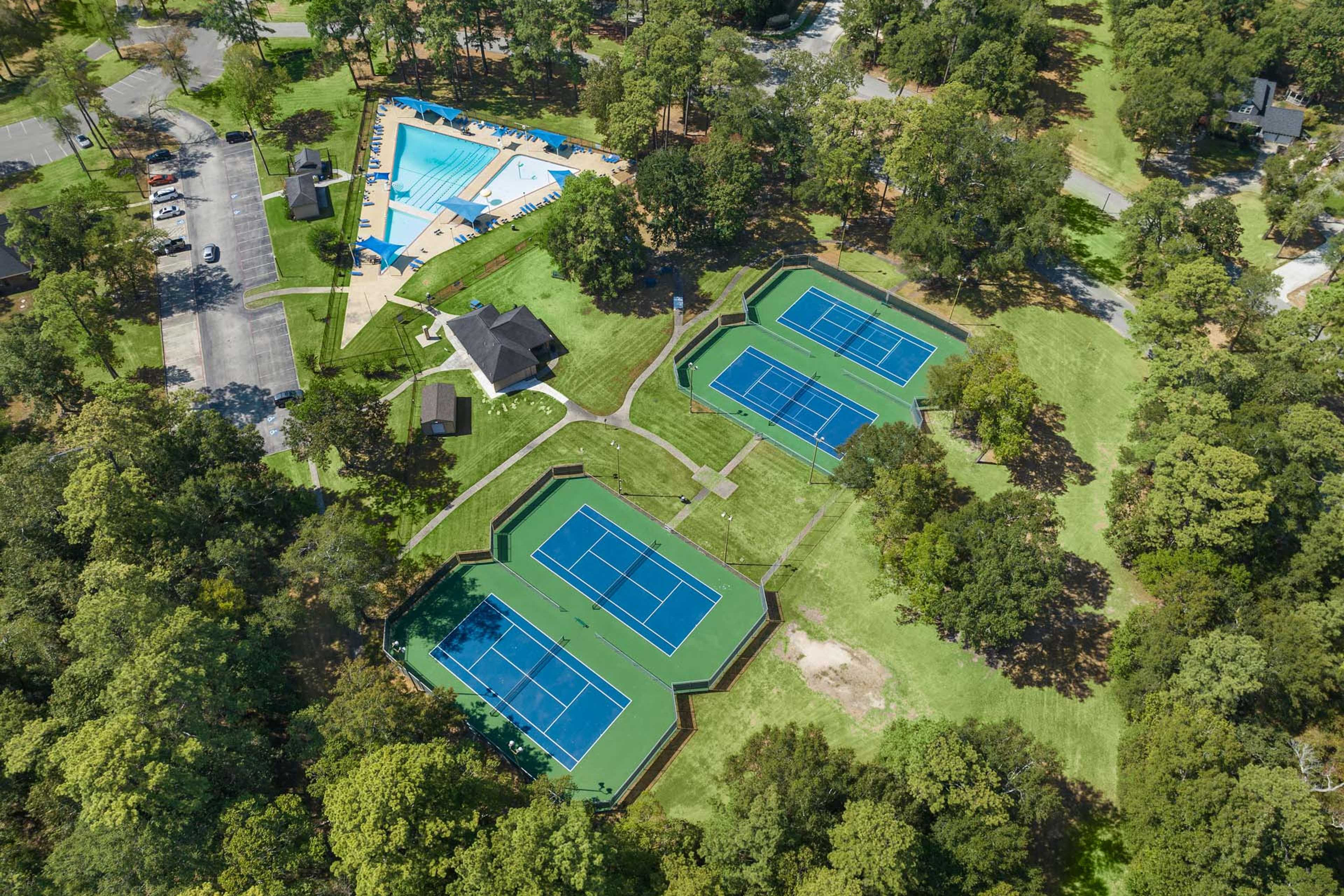 Aerial view of resort-style swimming pool and three blue tennis courts at Enclave at Newport in Crosby Texas surrounded by lush greenery