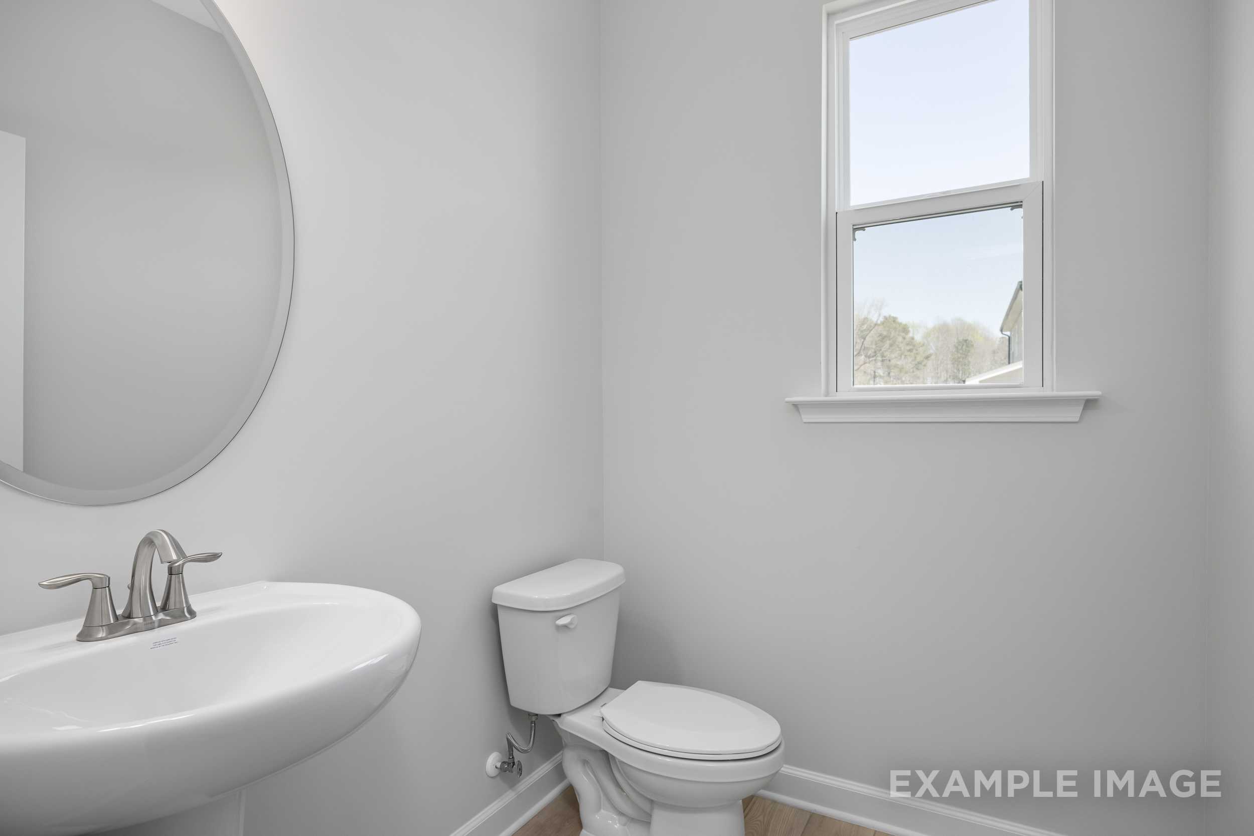 Elegant powder room in The Hickory II featuring round mirror, white pedestal sink, toilet, and window in soft gray walls