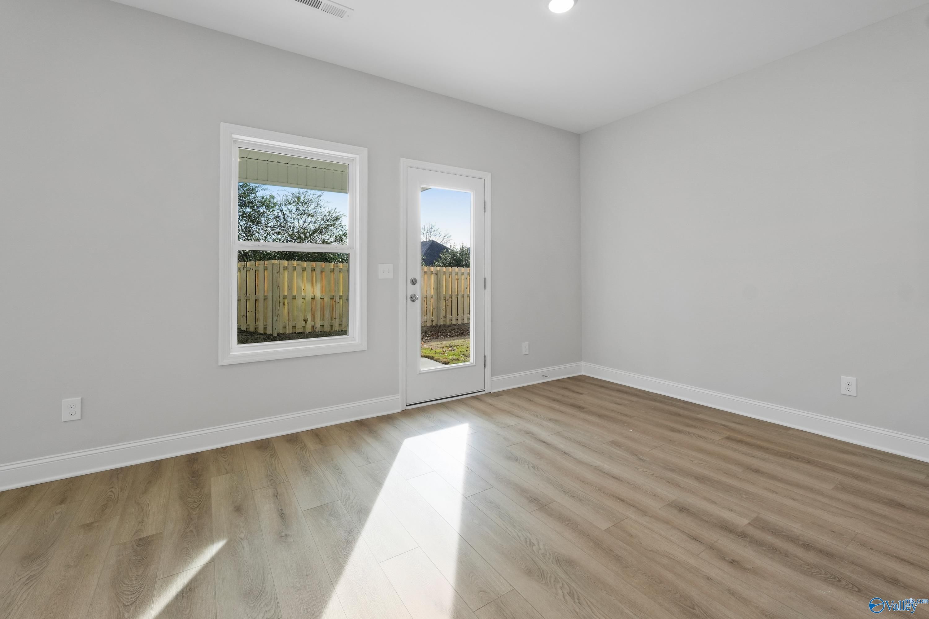 Sunlit empty room with gray walls, hardwood floors, large windows, and glass door to fenced backyard in Davidson Homes The Camden, Huntsville AL