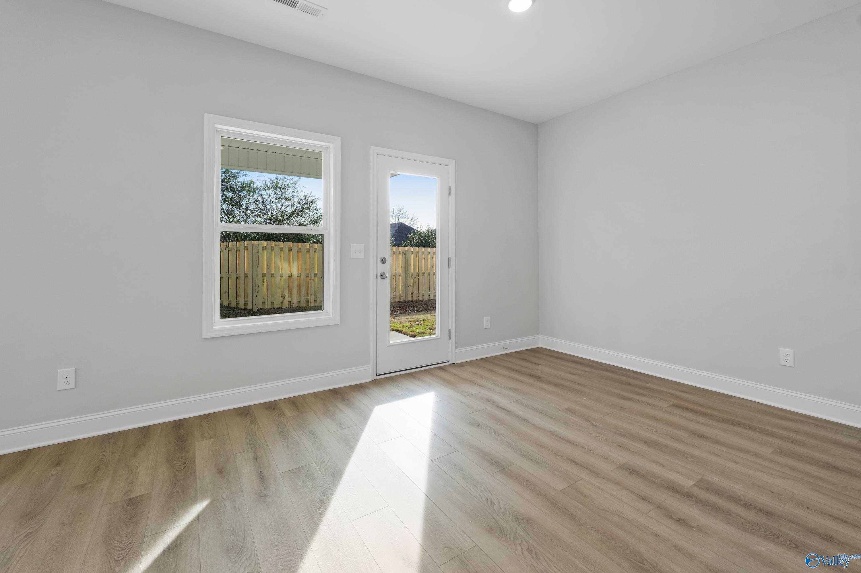 Sunlit empty room with gray walls, hardwood floors, large windows, and glass door to fenced backyard in Davidson Homes The Camden, Huntsville AL