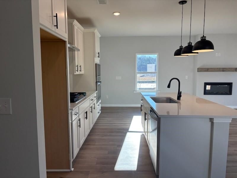 Modern open-concept kitchen with white shaker cabinets, quartz island sink, stainless dishwasher, and adjacent fireplace in Davidson Homes The Rabun C, Winder, GA