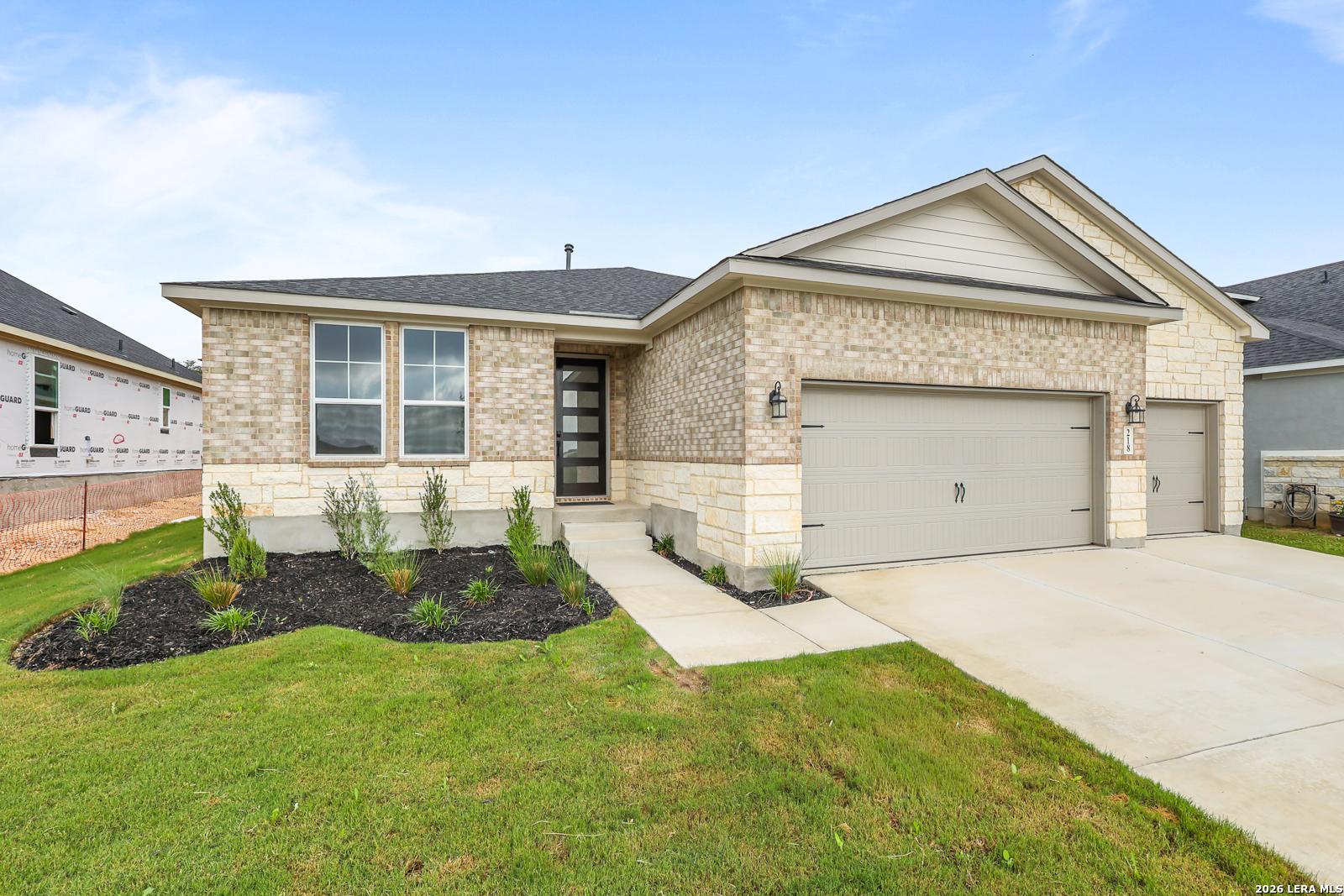 Modern tan brick 1-story home with 2-car garage, stone accents, and landscaped yard in The Reserve at Potranco Oaks, Castroville, Texas