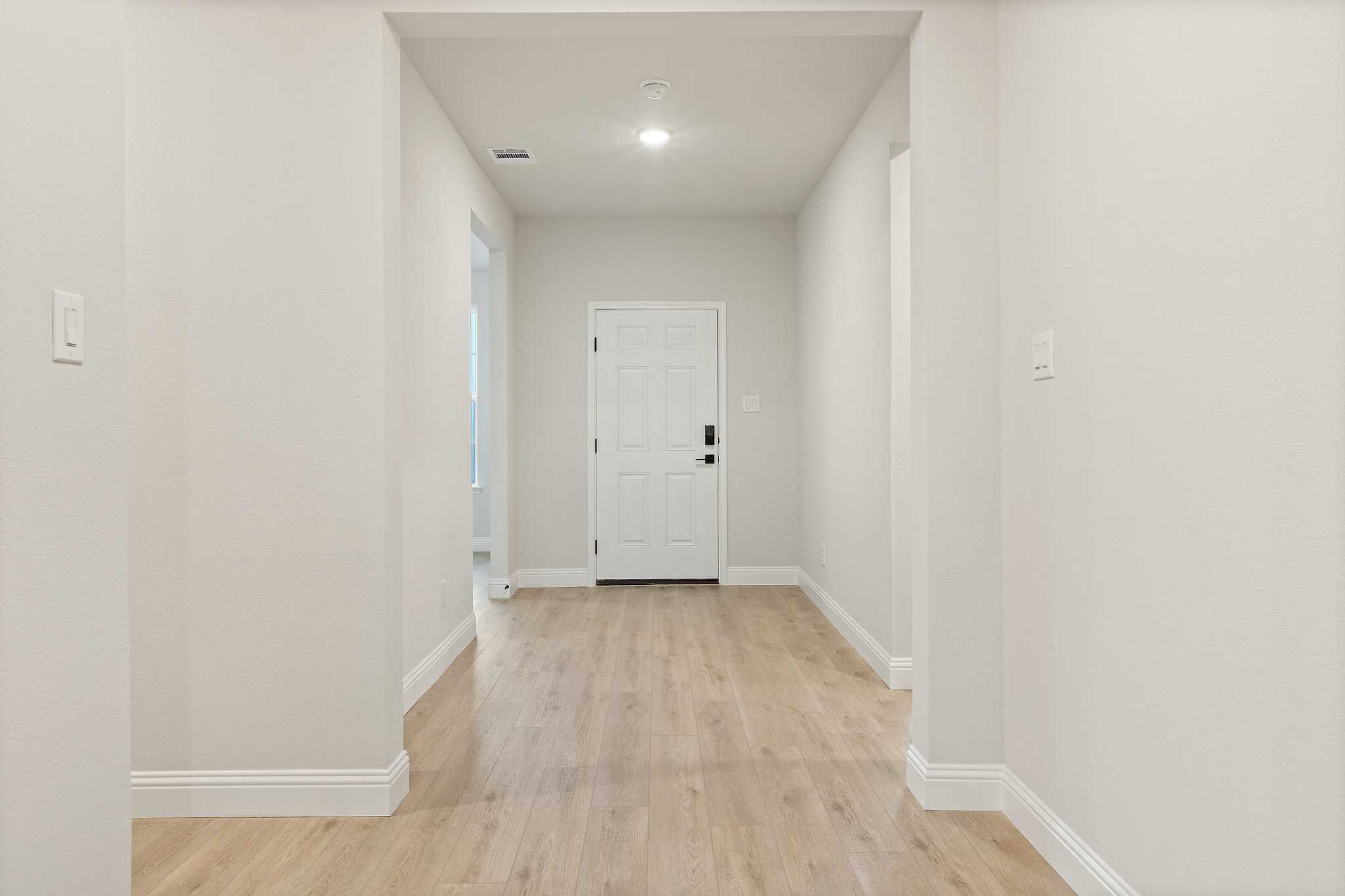 Spacious hallway in The Harrison C home featuring light oak floors, neutral walls, and white door with soft ceiling light