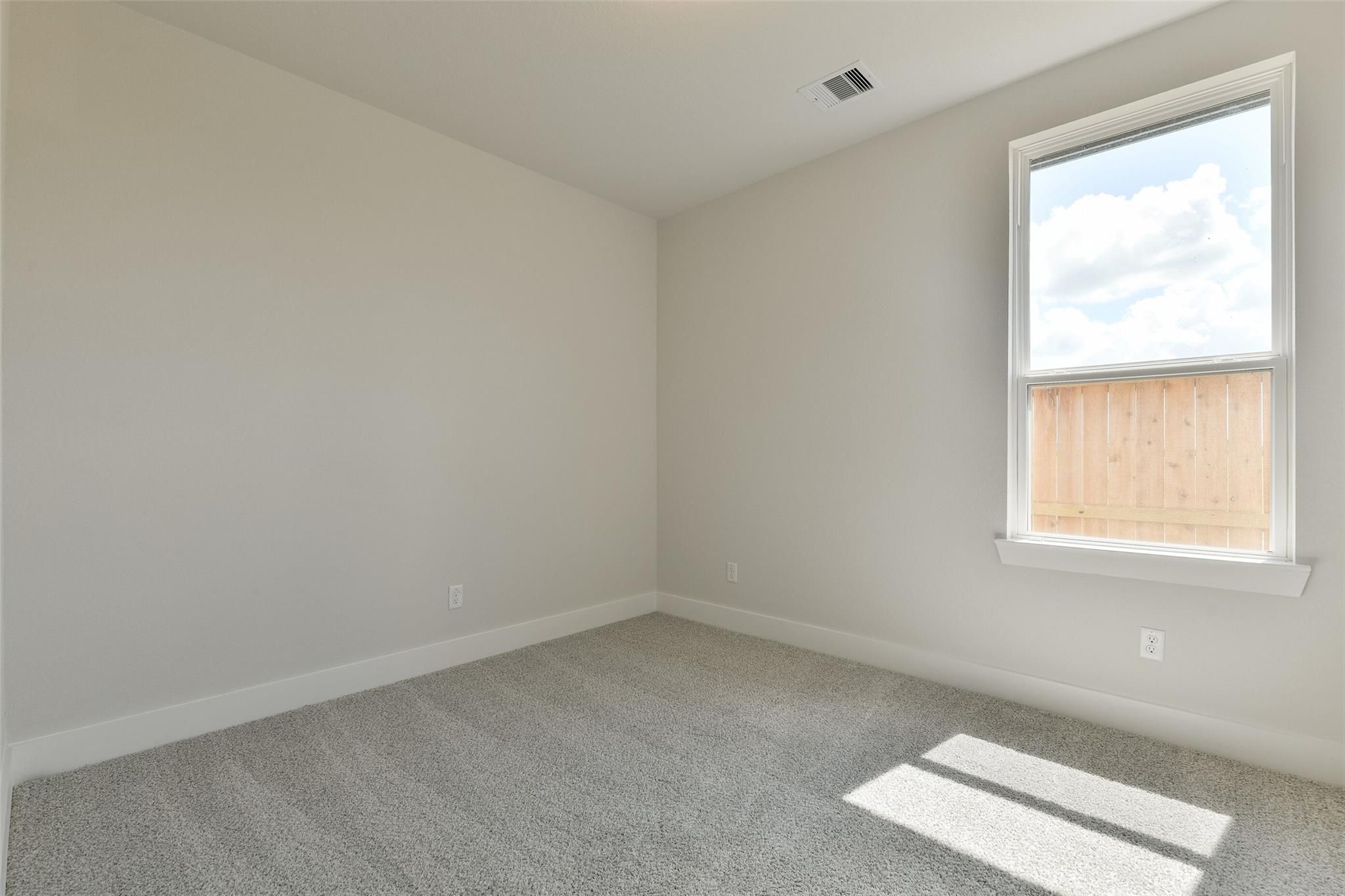 Bright secondary bedroom with beige walls, gray carpet, and window overlooking wooden fence in Davidson Homes The Edward A, Lago Mar, Texas