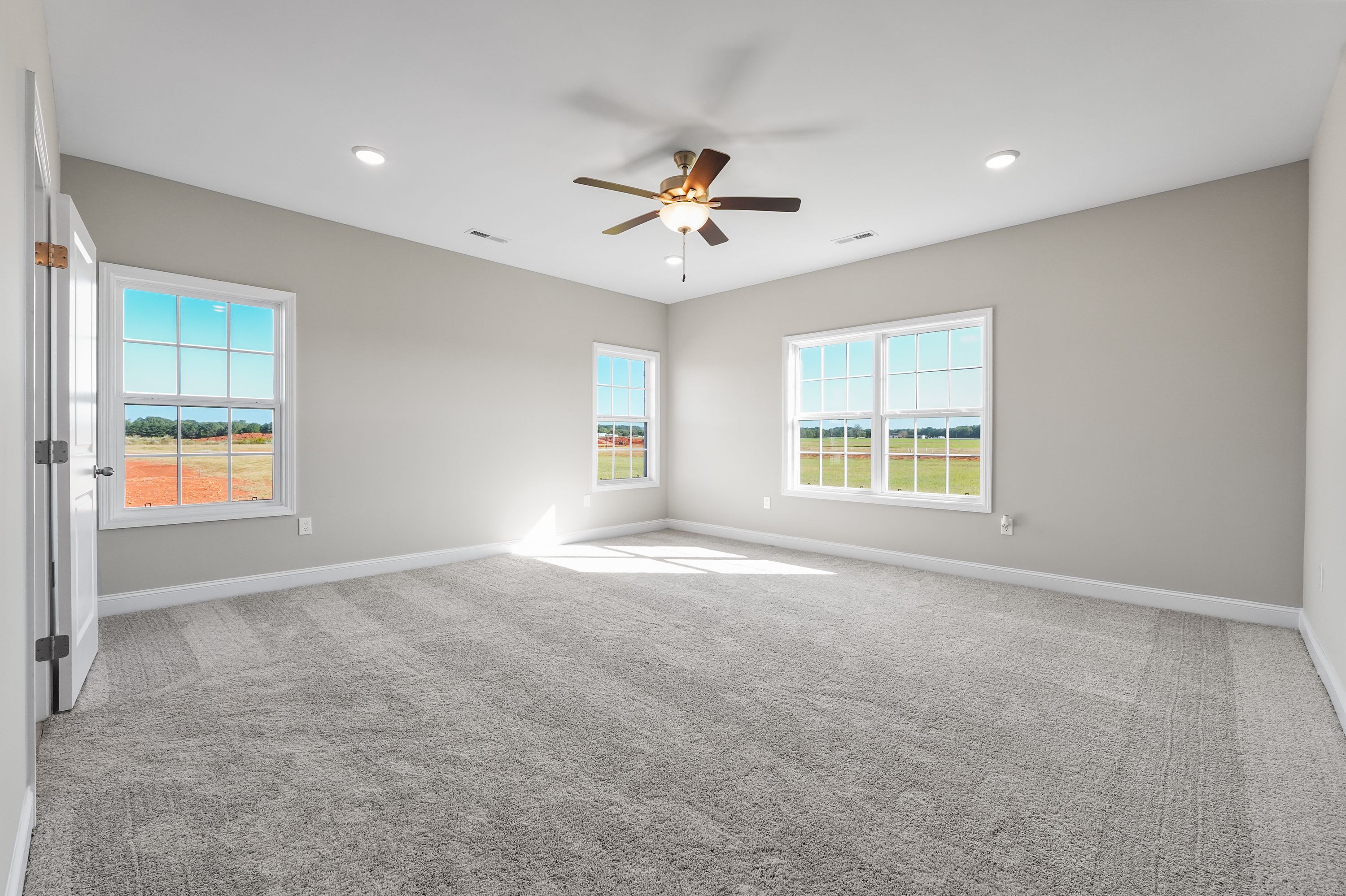 Spacious master bedroom in The Valencia home with gray walls, beige carpet, ceiling fan, and large windows overlooking Alabama fields