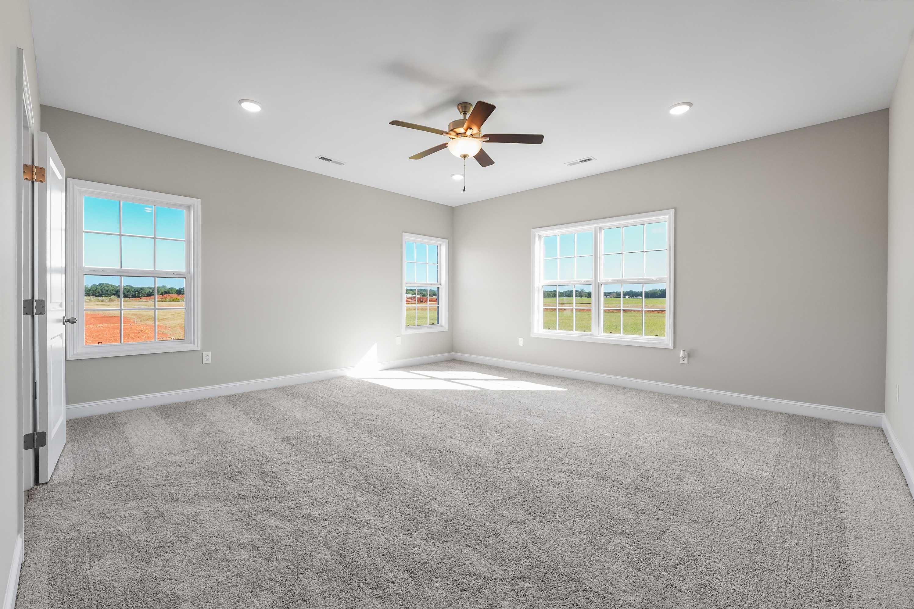 Spacious master bedroom in The Valencia home with gray walls, beige carpet, ceiling fan, and large windows overlooking Alabama fields