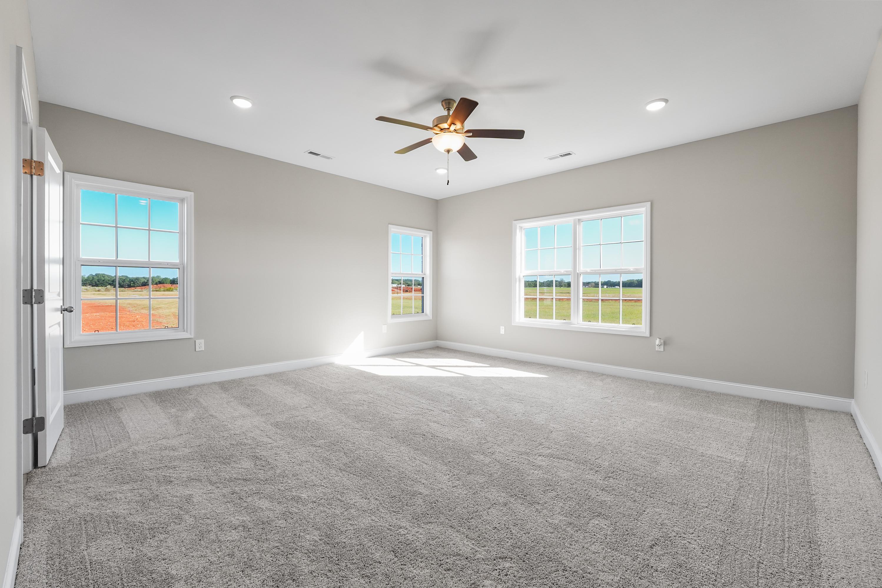 Spacious master bedroom in The Valencia home with gray walls, beige carpet, ceiling fan, and large windows overlooking Alabama fields