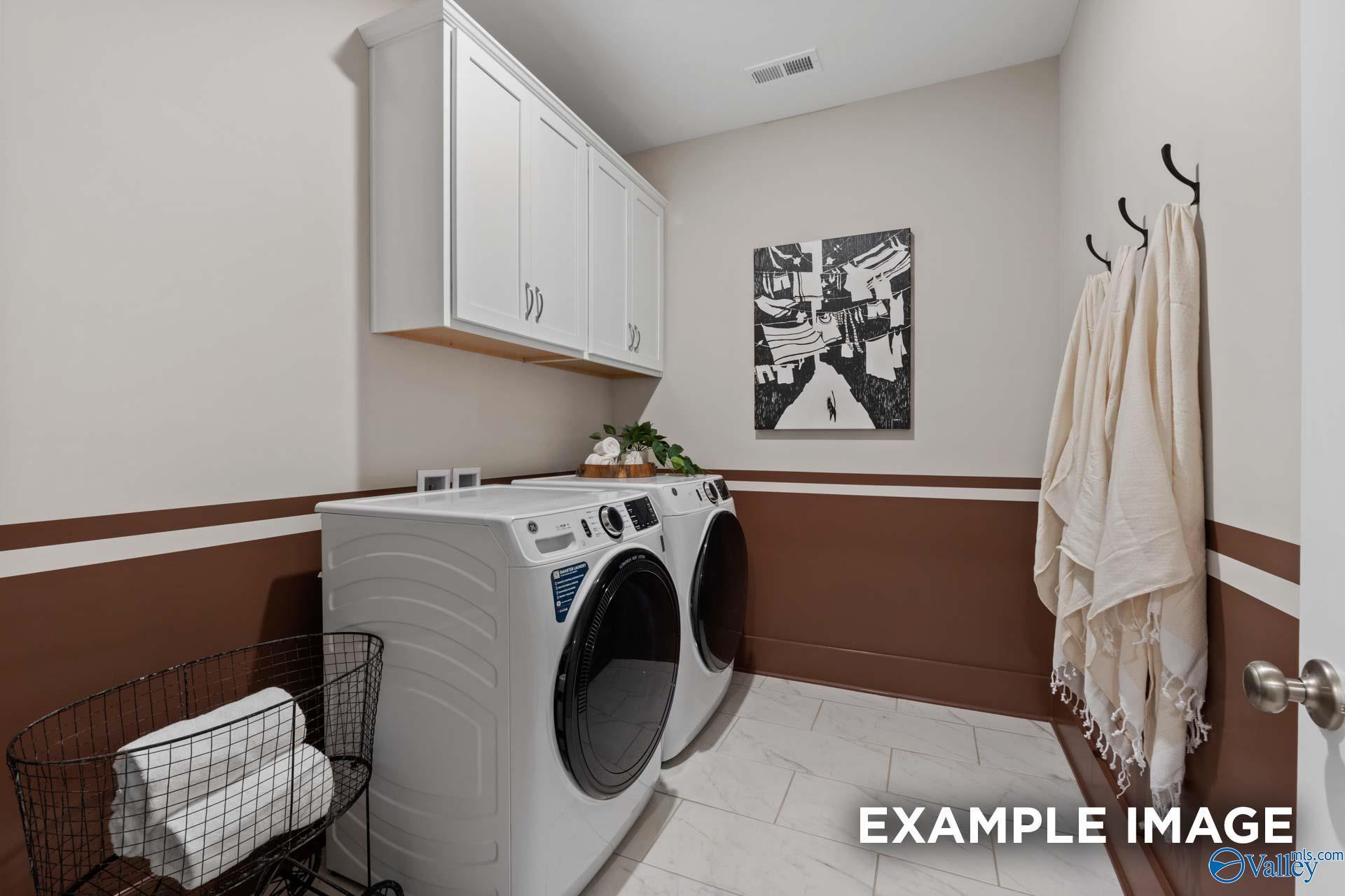Modern laundry room featuring white washer, dryer, cabinets, towel hooks, and abstract art in Davidson Homes The Finleigh, Huntsville, AL
