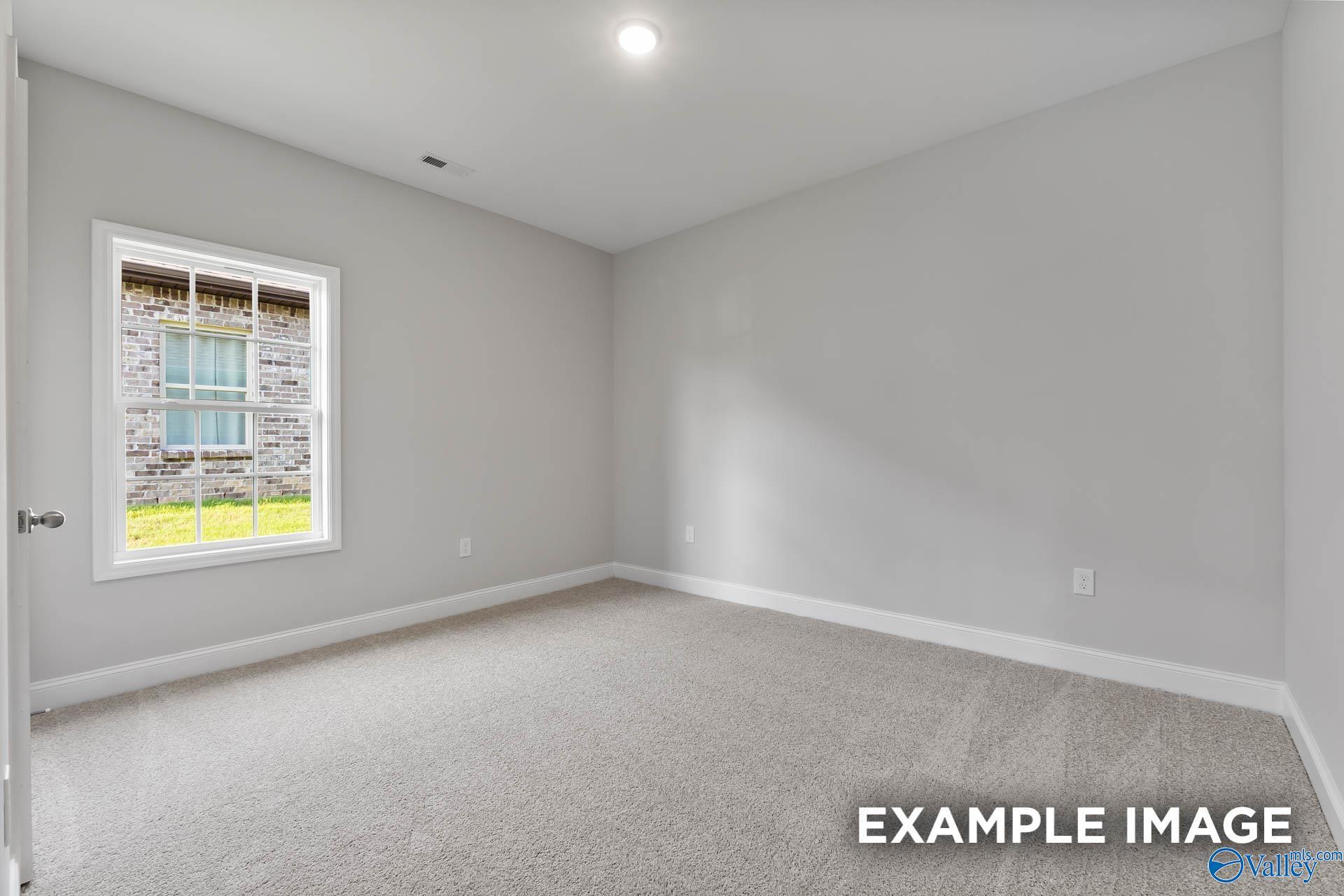 Bright empty bedroom with gray walls, beige carpet, ceiling light, and window view of brick exterior in The Daphne C home, Toney, Alabama
