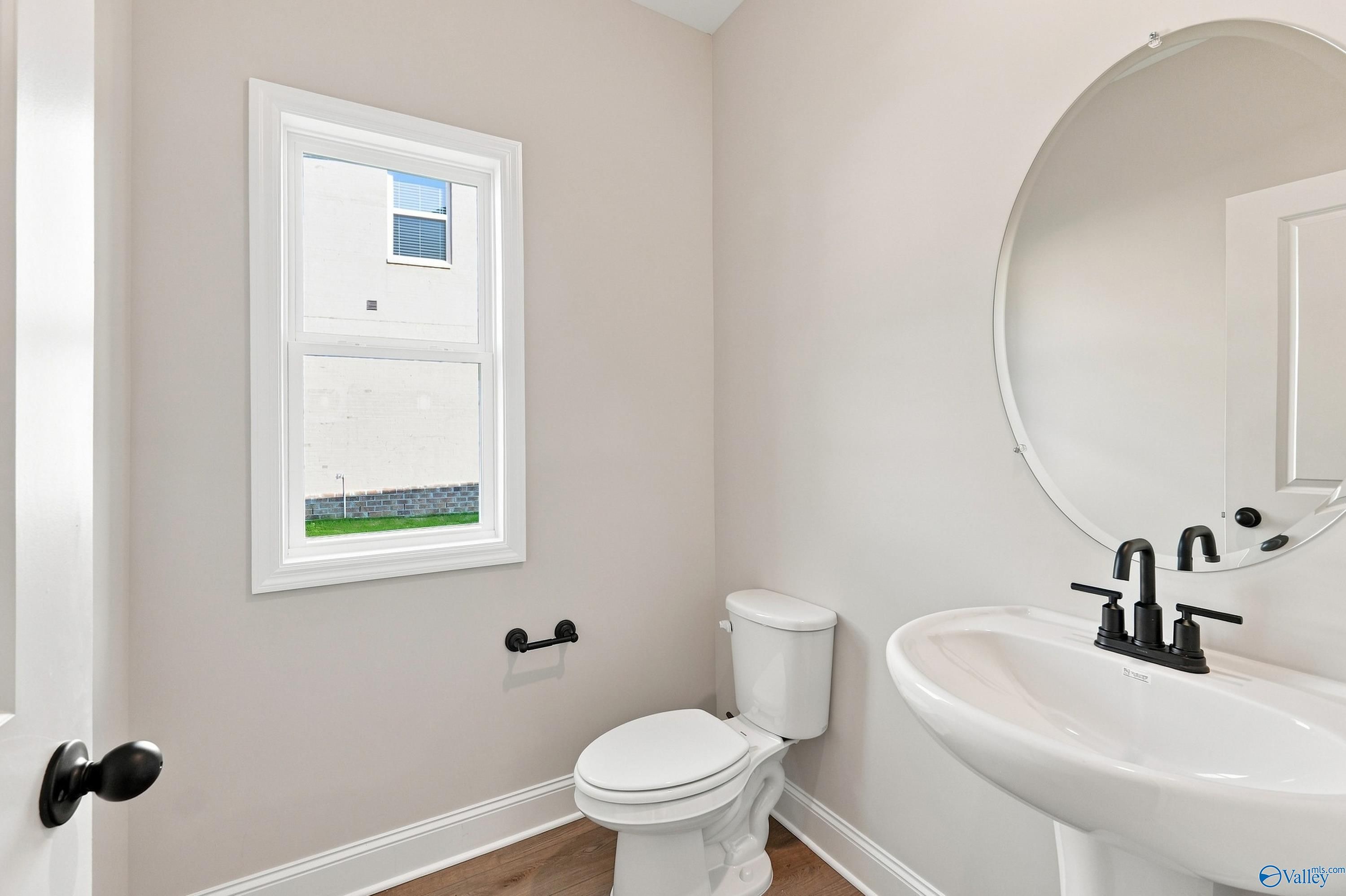 Bright powder room with pedestal sink, black faucet, round mirror, toilet, and window in Davidson Homes The Chelsea B, New Market, AL