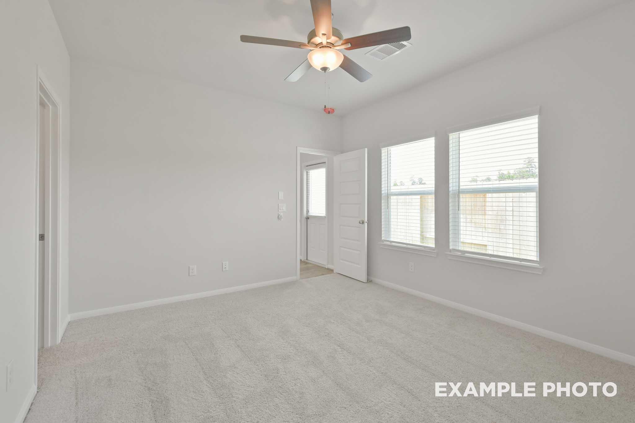 Spacious master bedroom in The Comal by Davidson Homes featuring ceiling fan, neutral gray walls, beige carpet, and large windows in Magnolia Texas