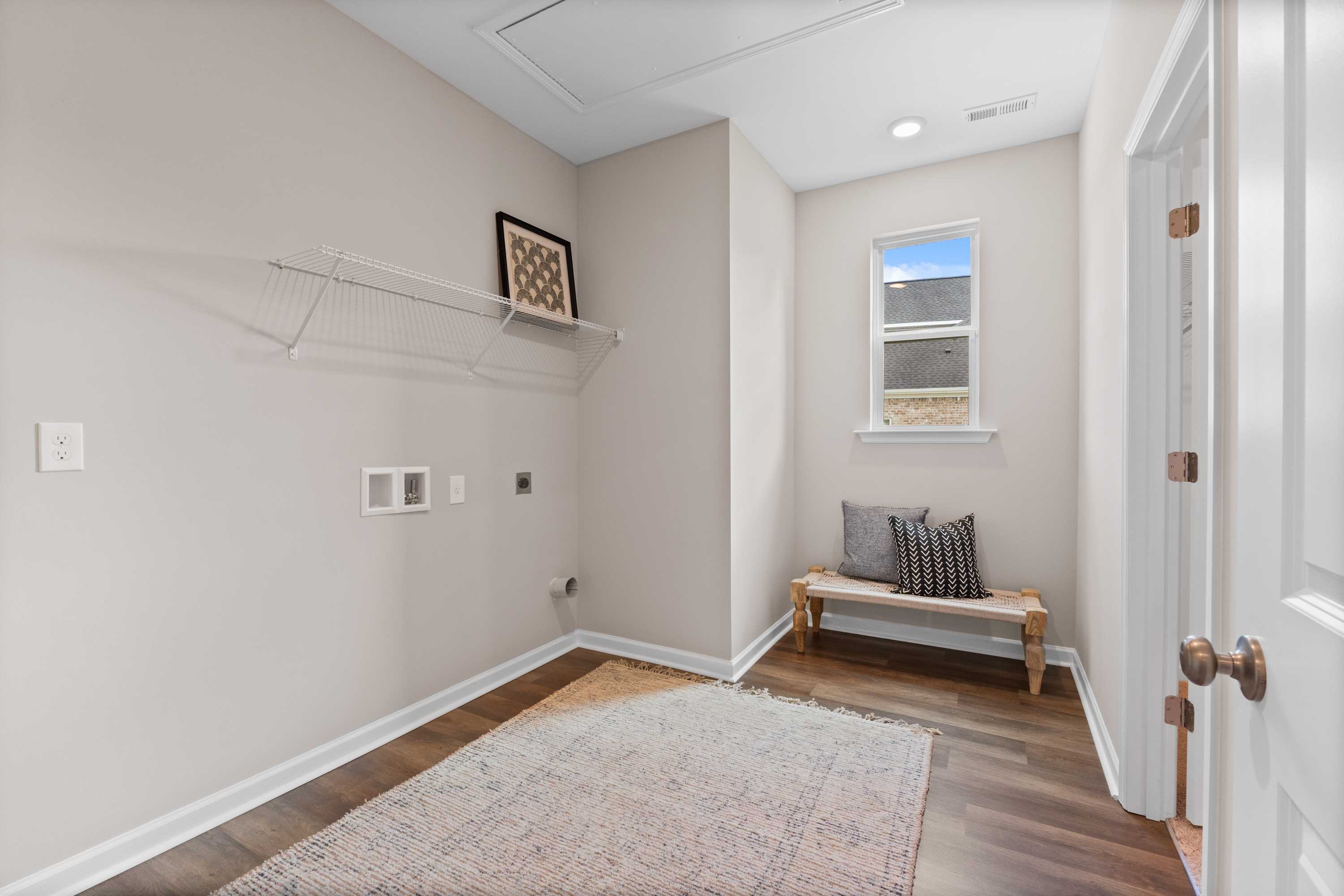 Spacious laundry room at Riverton Preserve in Huntsville AL featuring beige walls, hardwood floors, built-in bench, shelves, and window