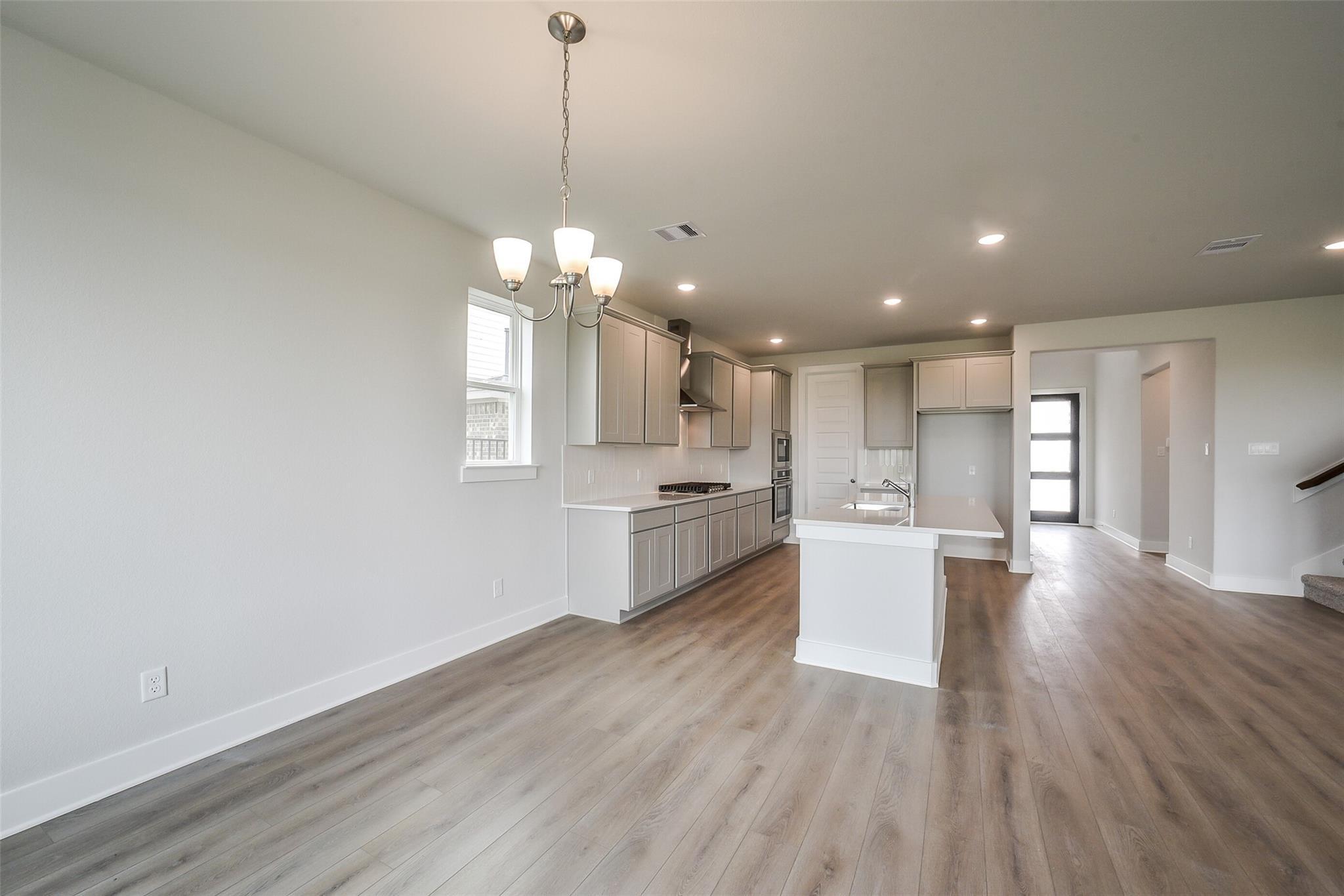 Modern open-concept kitchen with white cabinets, large island sink, stainless appliances, and wood flooring in Davidson Homes The Philip B, Rosharon, Texas