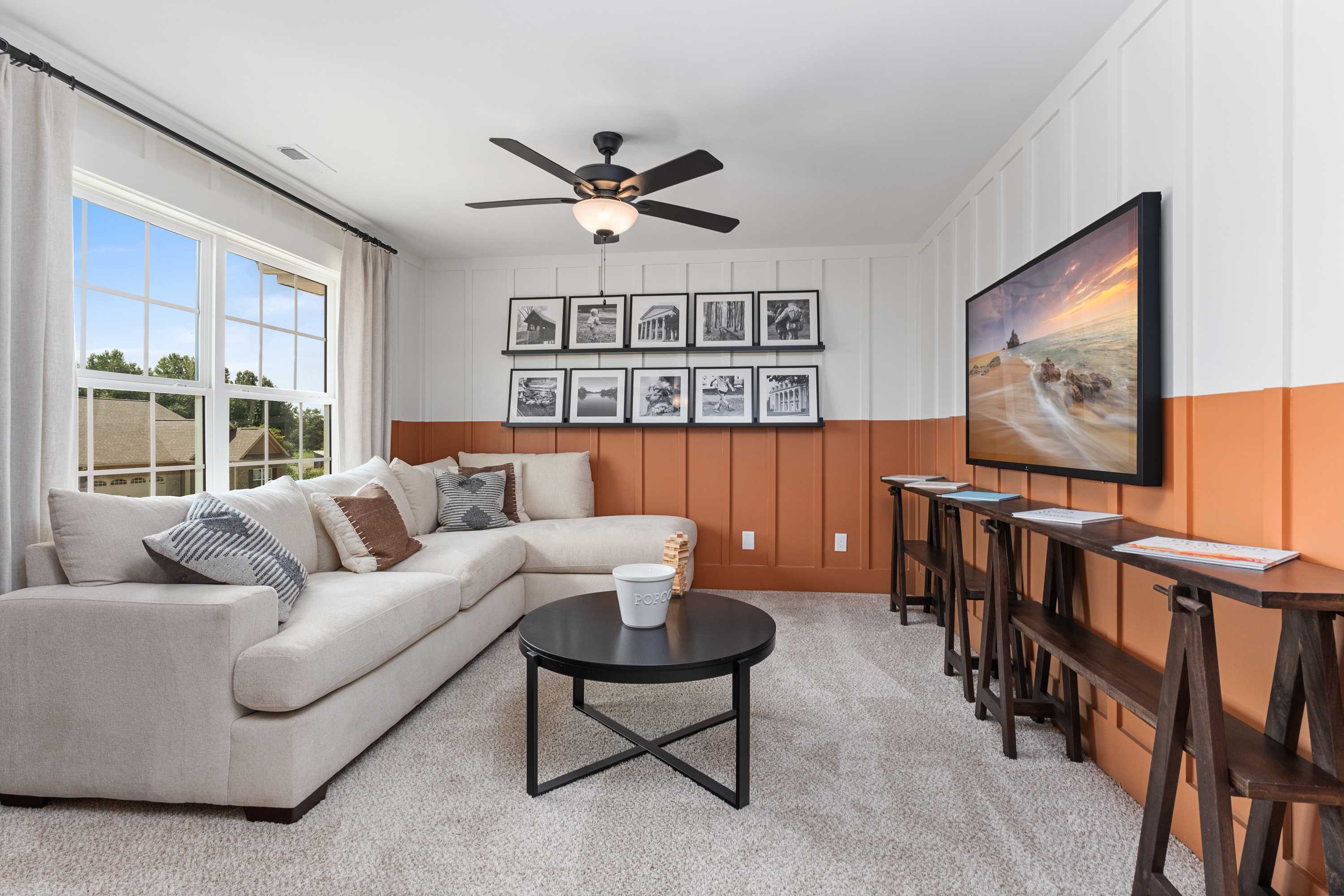 Cozy upstairs loft in The Shelby A featuring L-shaped beige sofa, wall-mounted TV, ceiling fan, and shiplap walls with orange accents