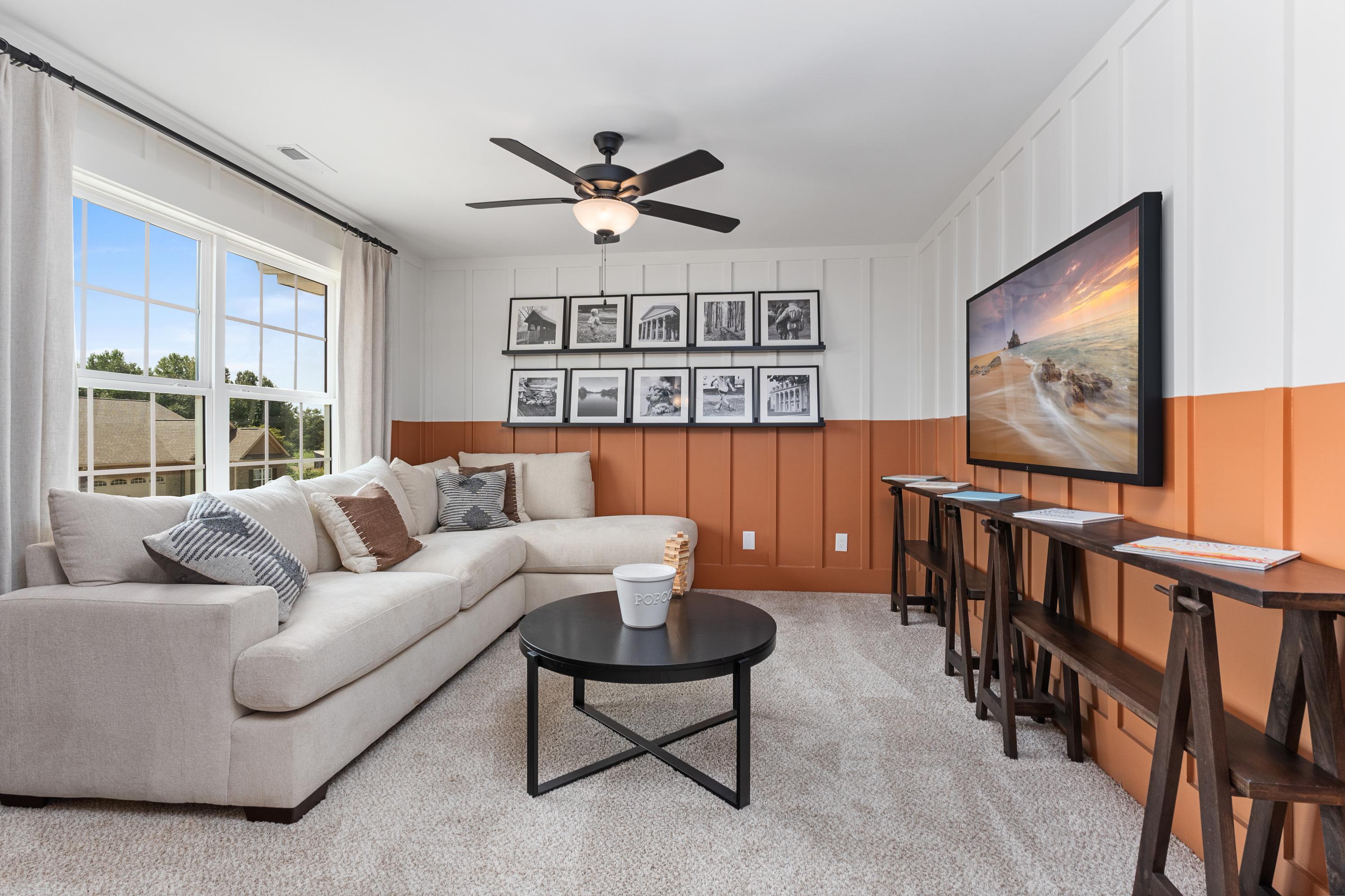 Cozy upstairs loft in The Shelby A featuring L-shaped beige sofa, wall-mounted TV, ceiling fan, and shiplap walls with orange accents