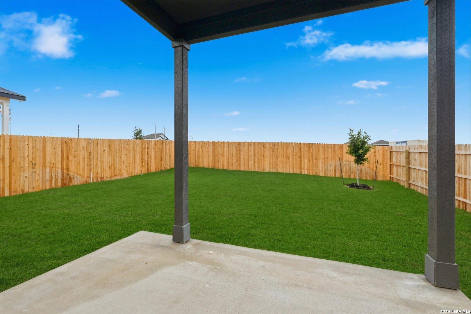 Covered back patio with concrete slab overlooking lush green backyard, wooden fence, and young tree in Davidson Homes The Blanco C, Agave, San Antonio