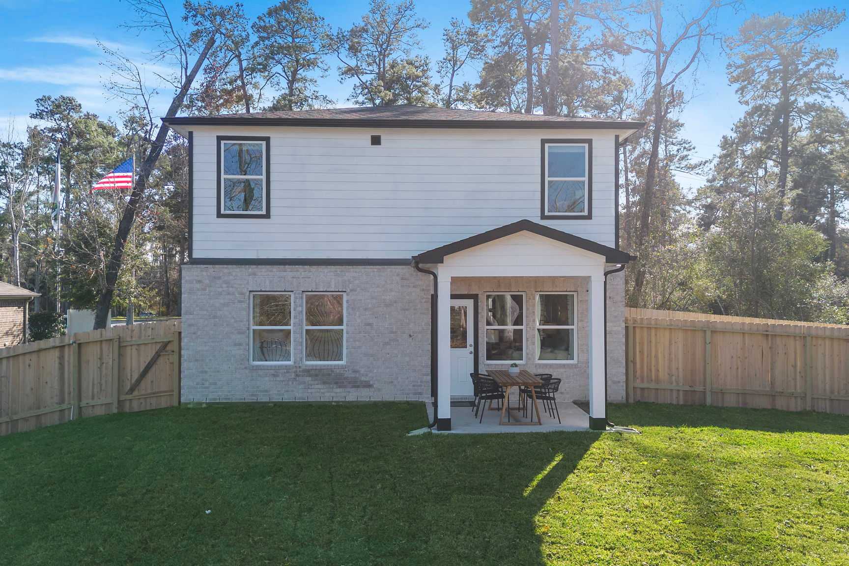 Rear view of modern two-story home at Lakes at Black Oak in Magnolia Texas with covered back porch, patio seating, brick accents, and fenced green yard
