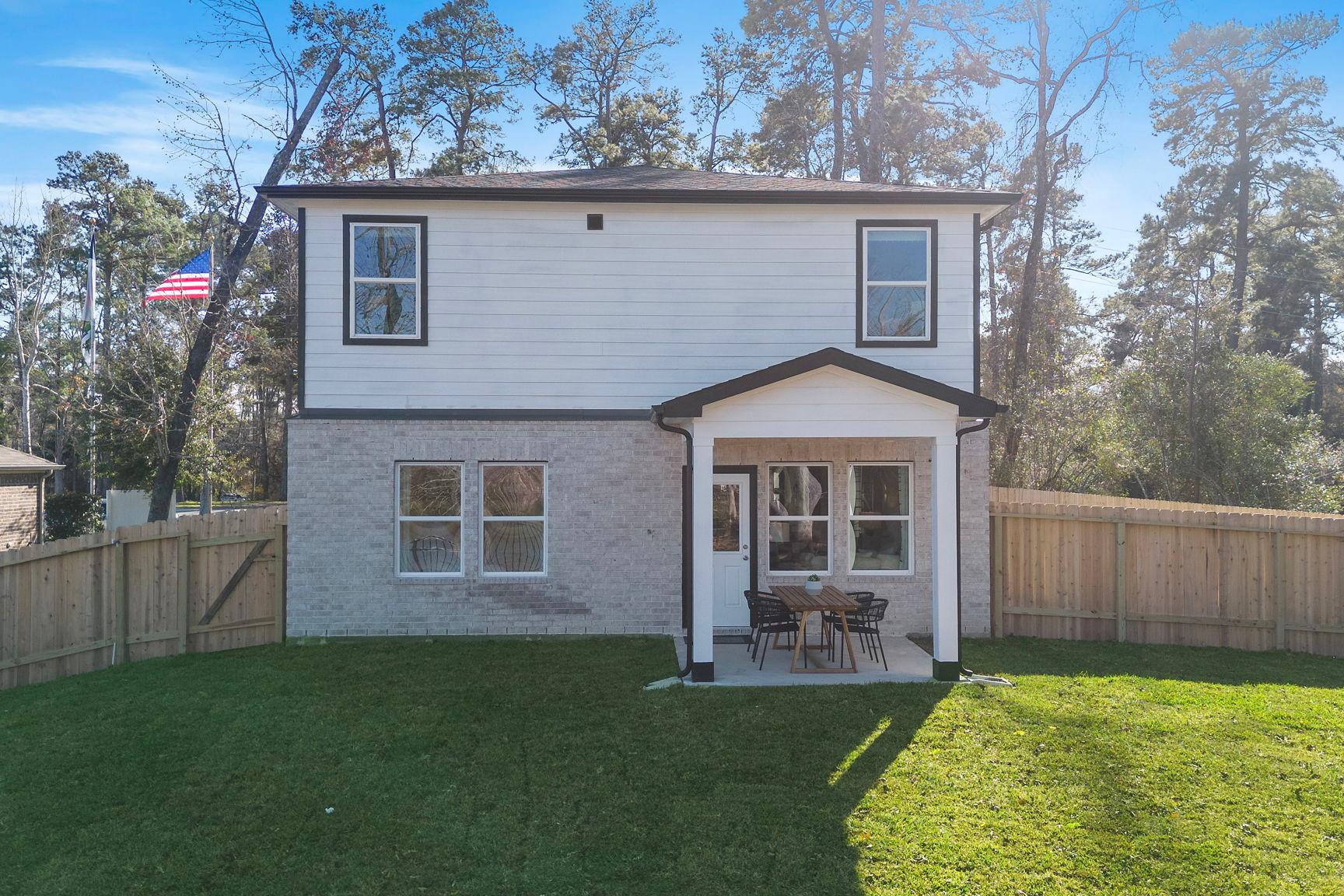 Rear view of modern two-story home at Lakes at Black Oak in Magnolia Texas with covered back porch, patio seating, brick accents, and fenced green yard