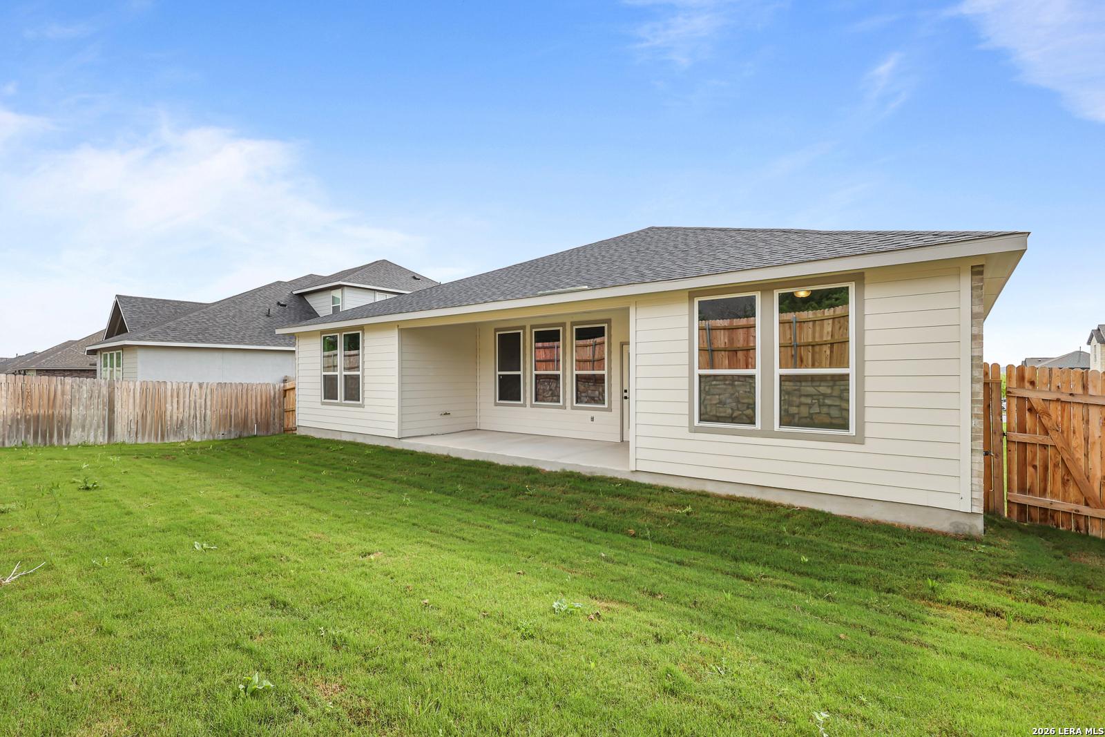 Single-story beige home with covered patio, large windows, and fenced green backyard in The Reserve at Potranco Oaks, Castroville, Texas