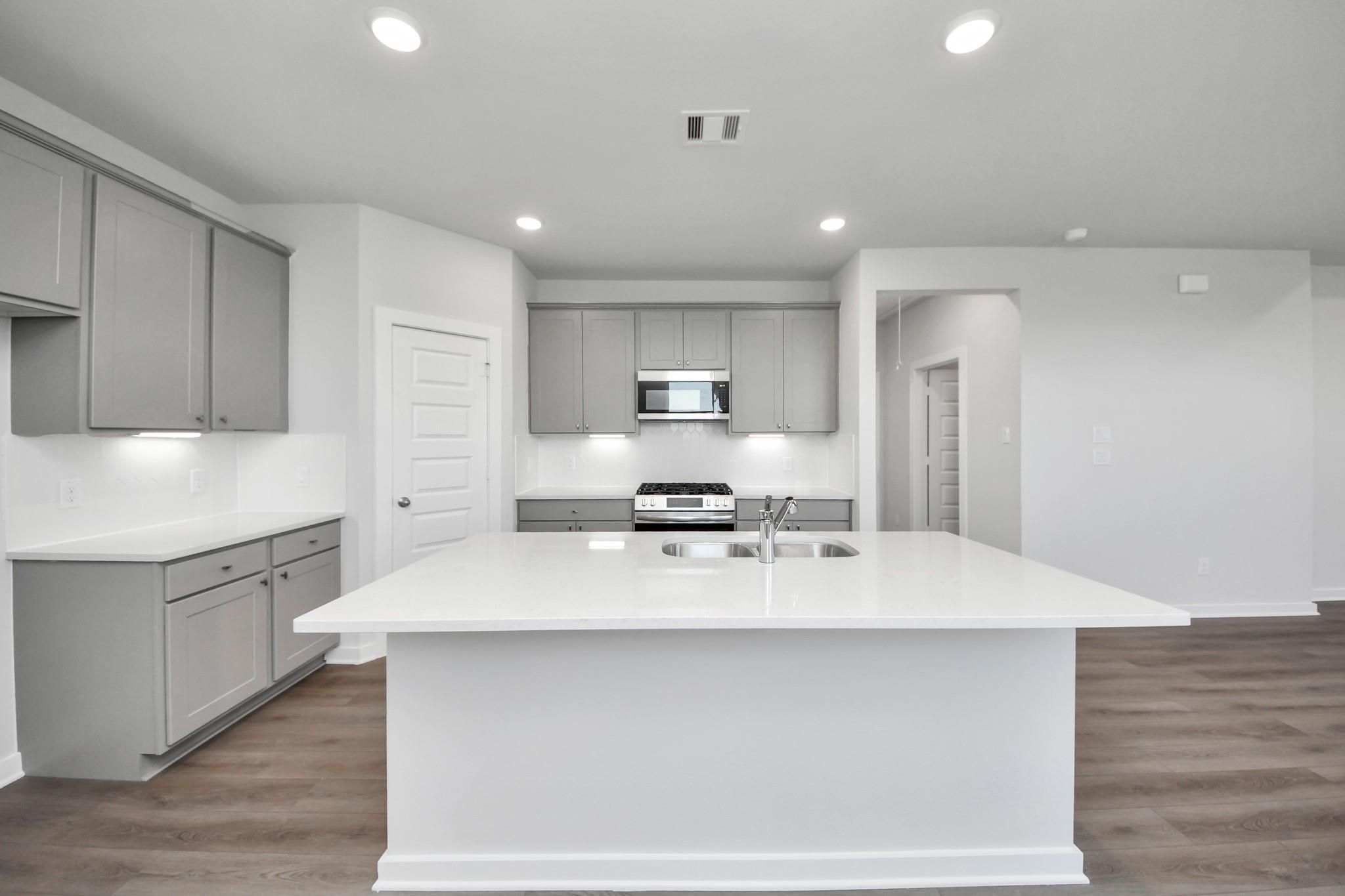 Modern kitchen with white island, gray cabinets, and stainless appliances in The Riviera A plan at Emberly in Beasley, TX.