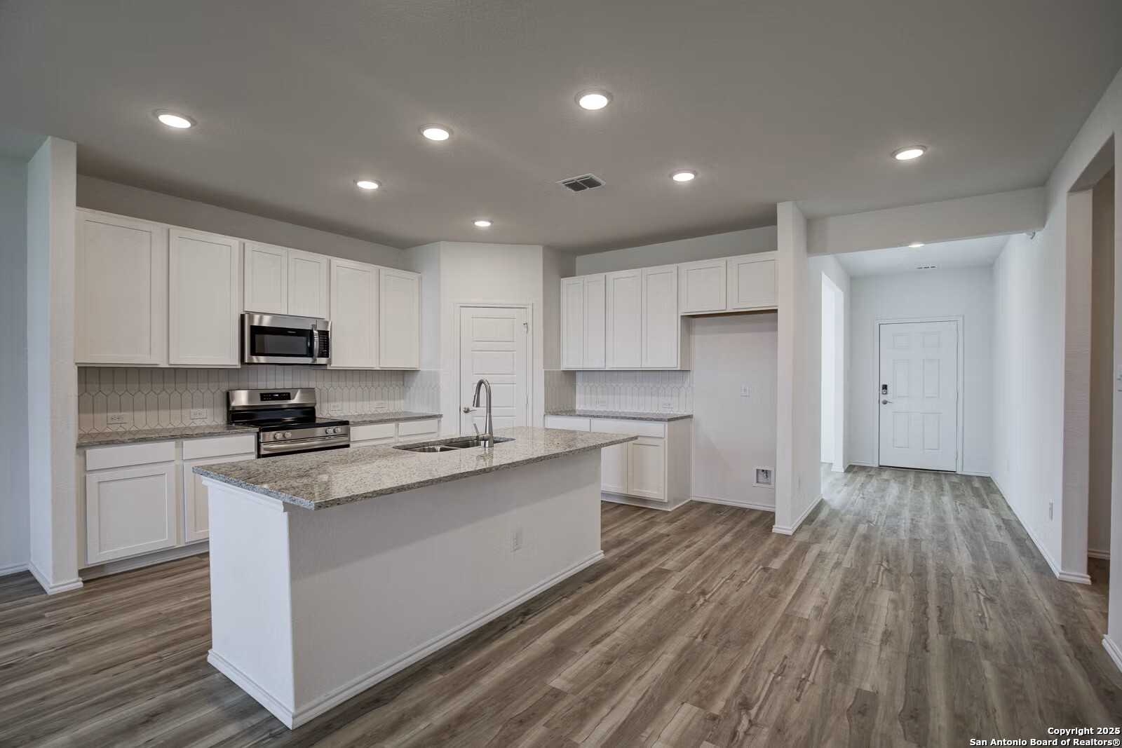 Modern white kitchen featuring granite island, stainless steel appliances, and wood flooring in Davidson Homes The Douglas B, Seguin, Texas