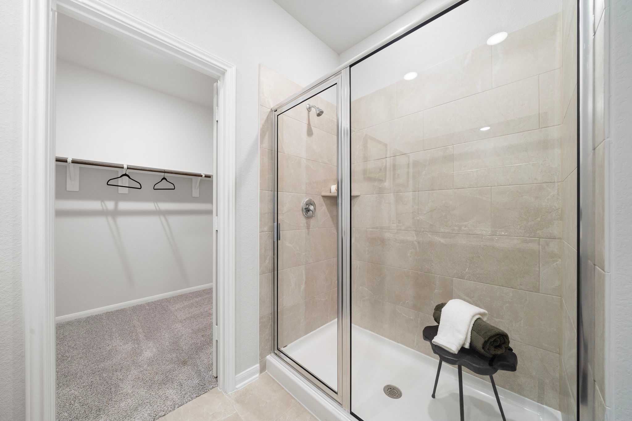 Spacious walk-in closet with shelving and hangers adjacent to modern glass shower enclosure, beige tile walls, and bench in Caney Creek Place Conroe Texas home