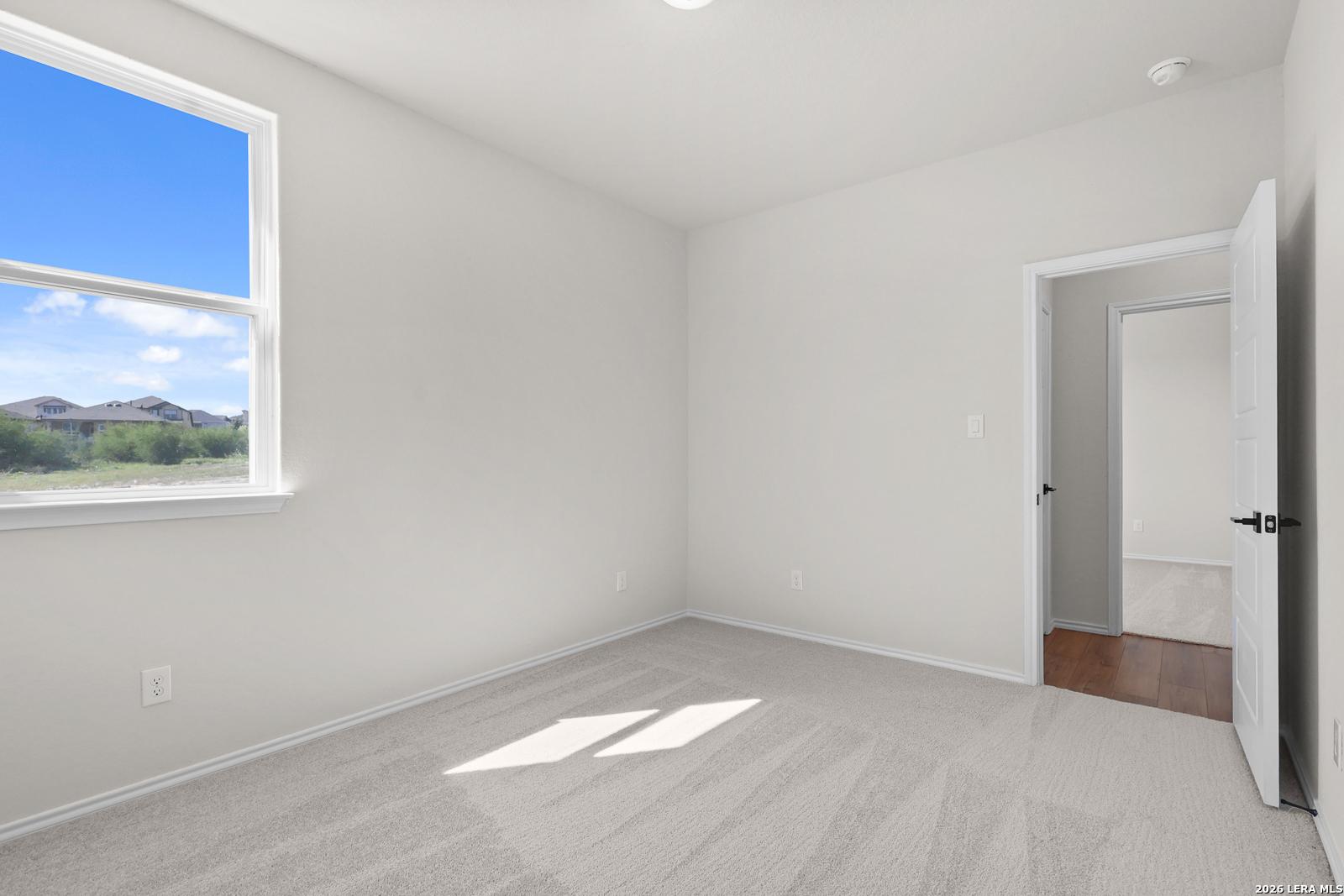 Bright empty bedroom with plush carpet, large window, and hardwood hallway in Davidson Homes Glenwood E, San Antonio, TX