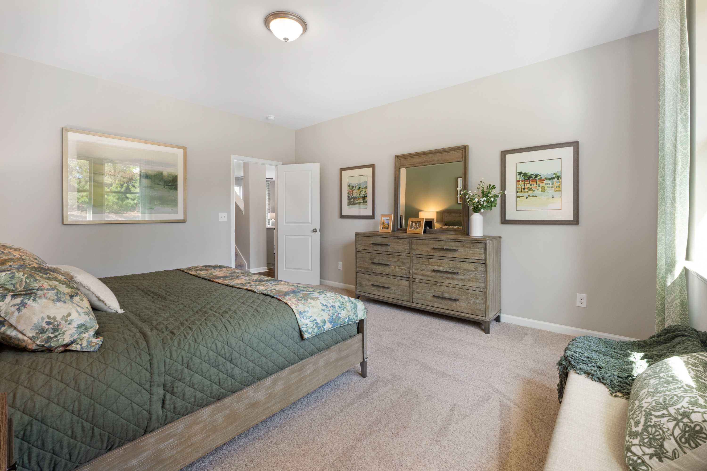 Spacious bedroom at Evergreen Mill in Madison, AL with wooden bed frame, green floral bedding, mirrored dresser, and wall art