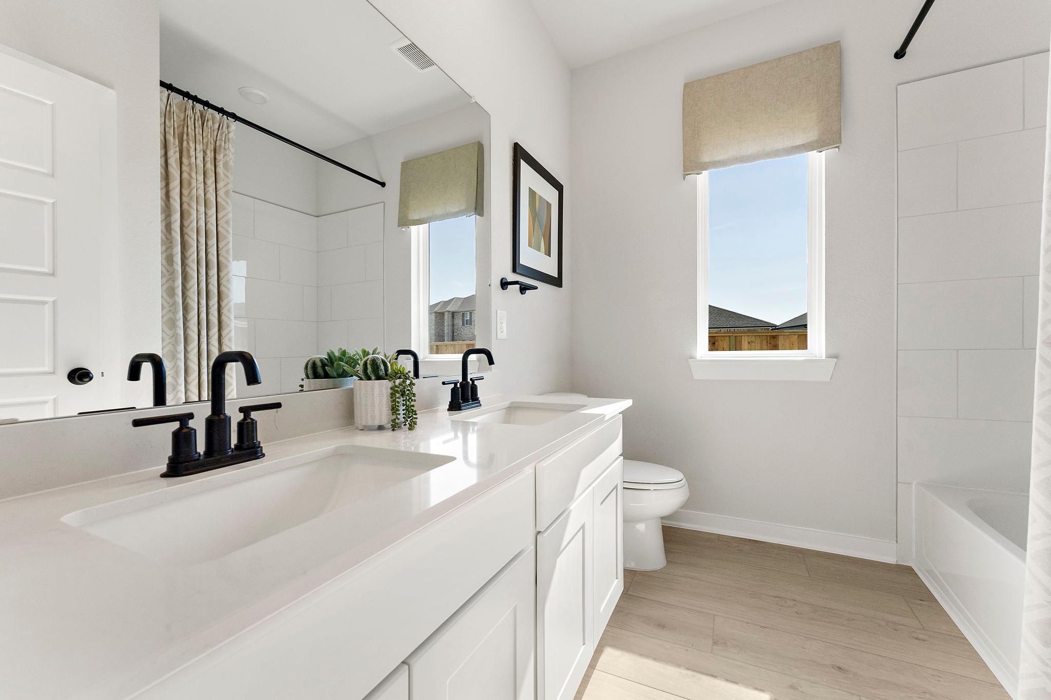 Modern white bathroom in Sundance Cove, Crosby Texas with double vanity, black faucets, bathtub, and light wood floors