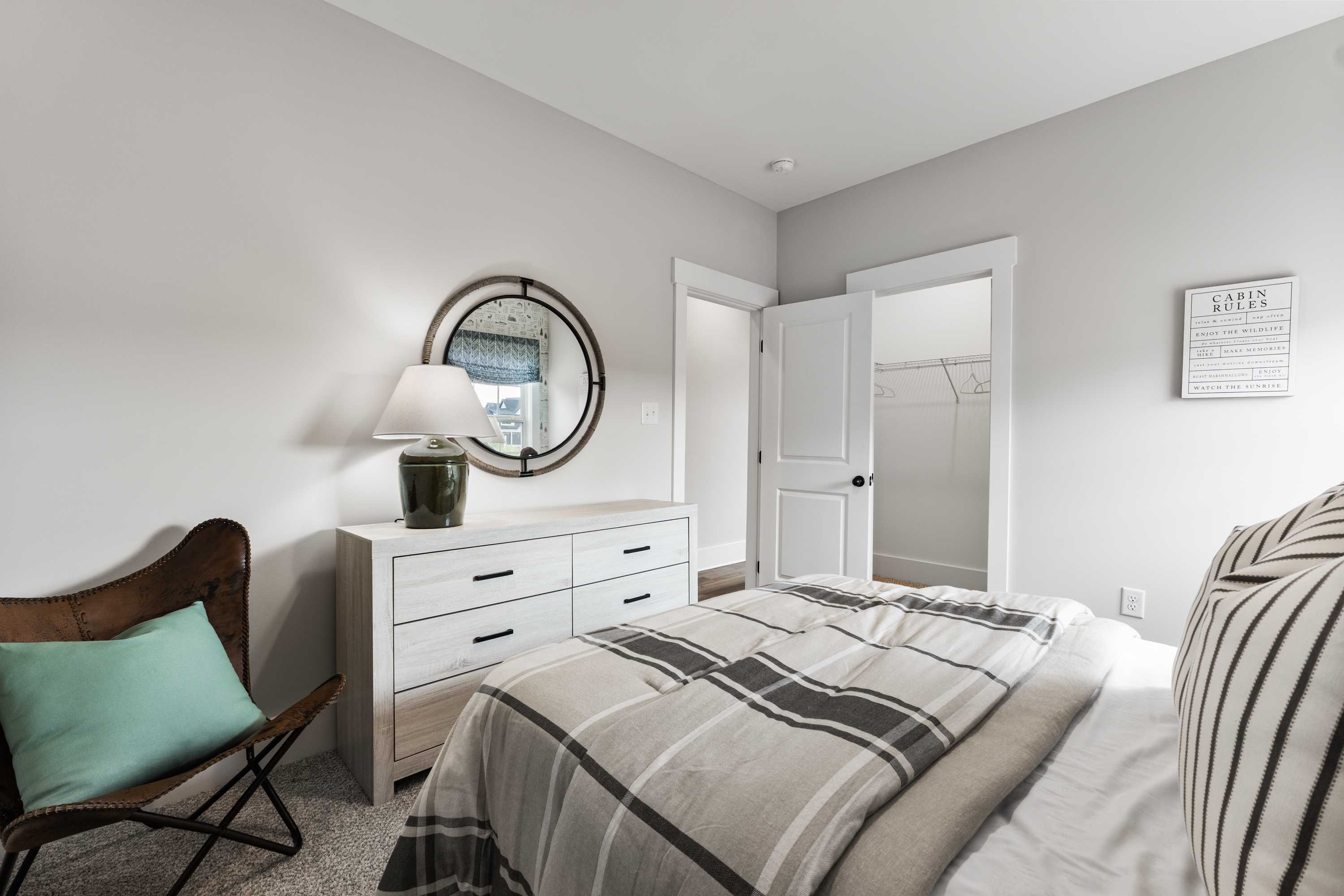 Cozy bedroom interior at River Road Estates in Decatur, Alabama with white dresser, round mirror, plaid bedding, and leather chair