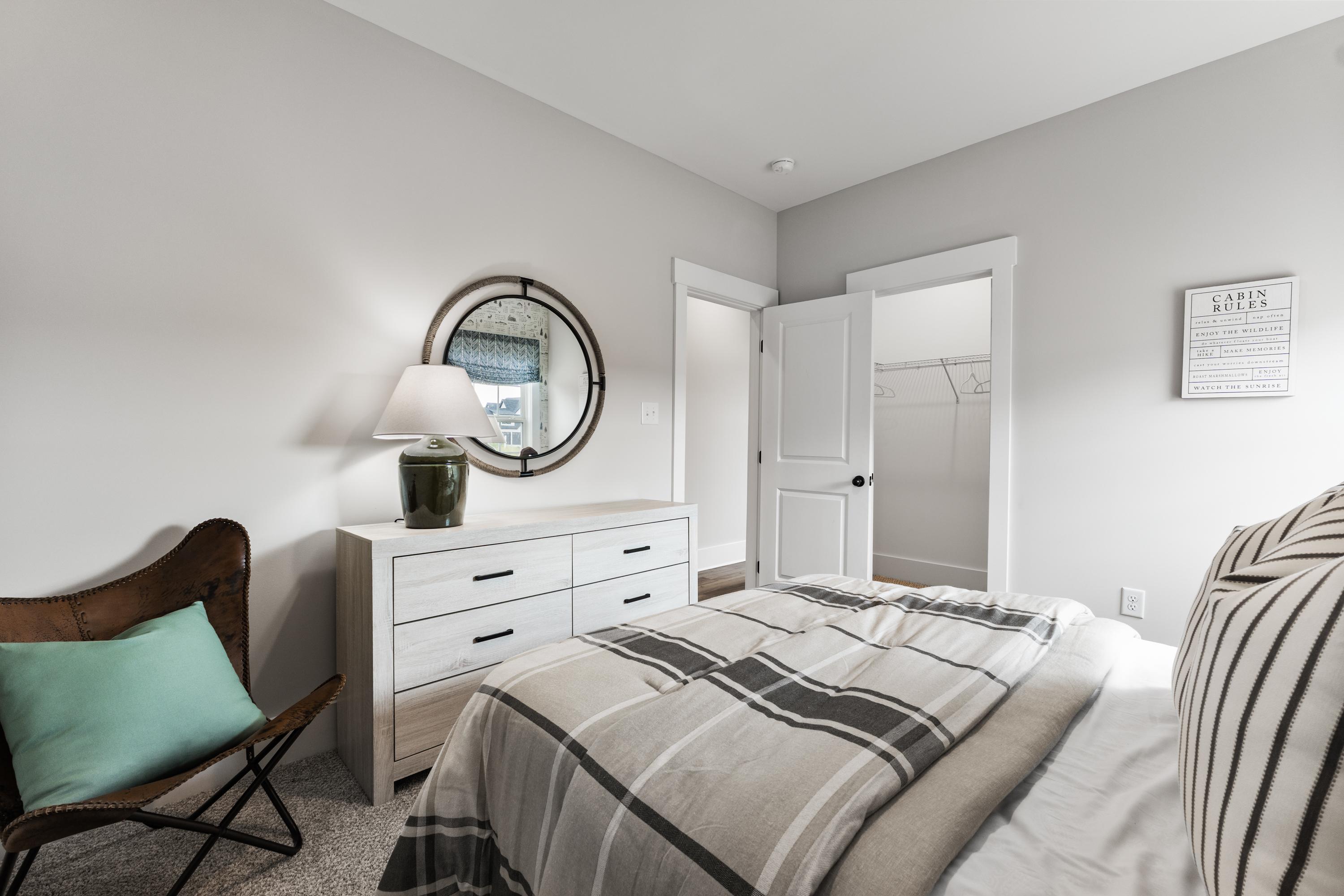 Cozy bedroom interior at River Road Estates in Decatur, Alabama with white dresser, round mirror, plaid bedding, and leather chair