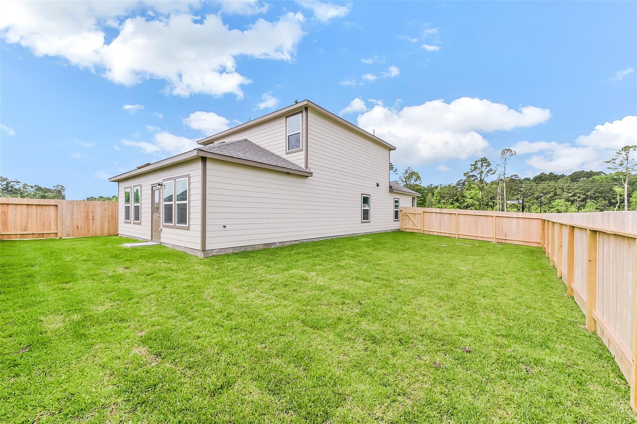 Two-story Sabine F home with beige siding, garage, lush green yard, wooden privacy fence, and wooded backdrop in Dayton, Texas