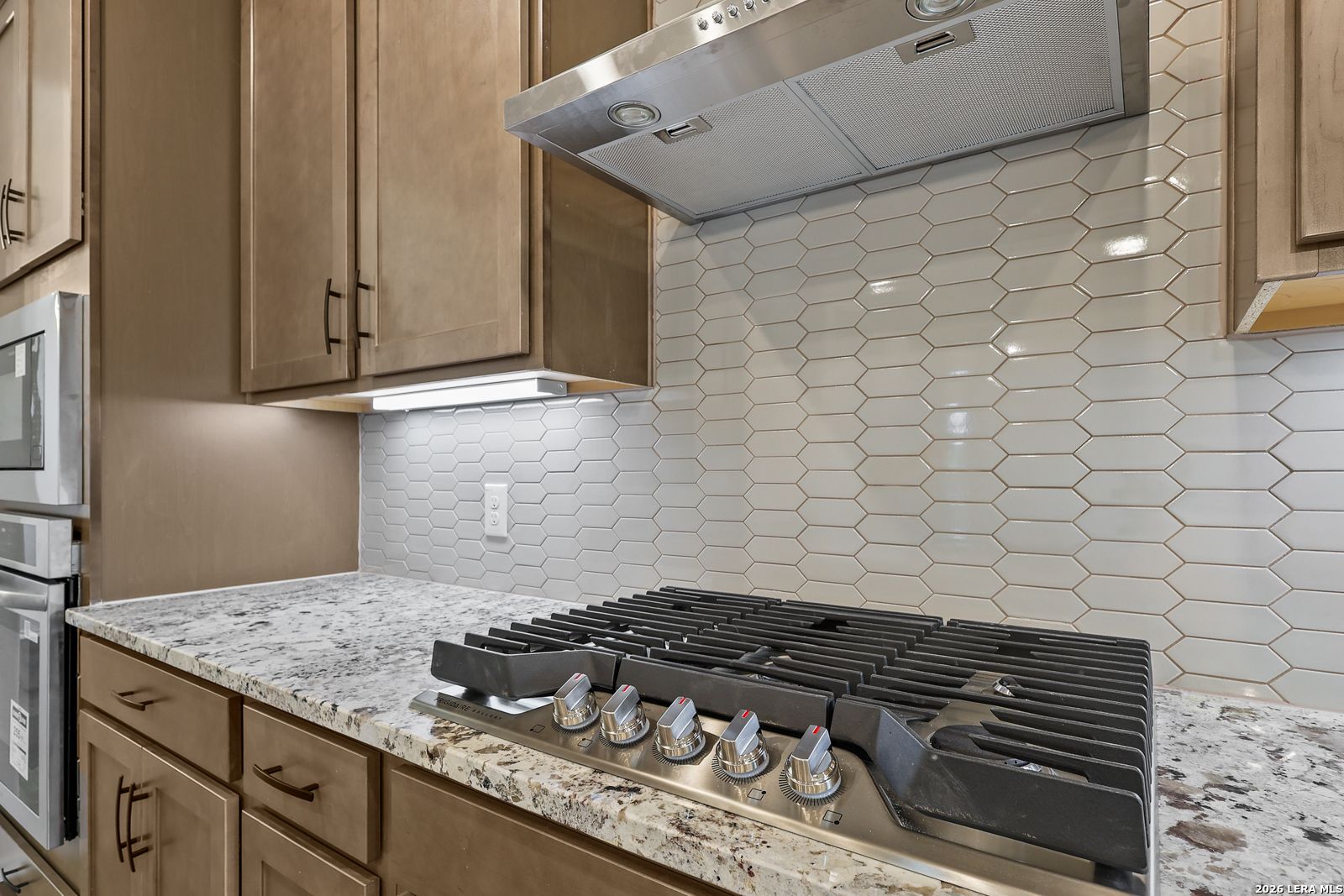 Gas cooktop with stainless hood, hexagon tile backsplash, and granite countertops in Davidson Homes Glenwood E kitchen, San Antonio