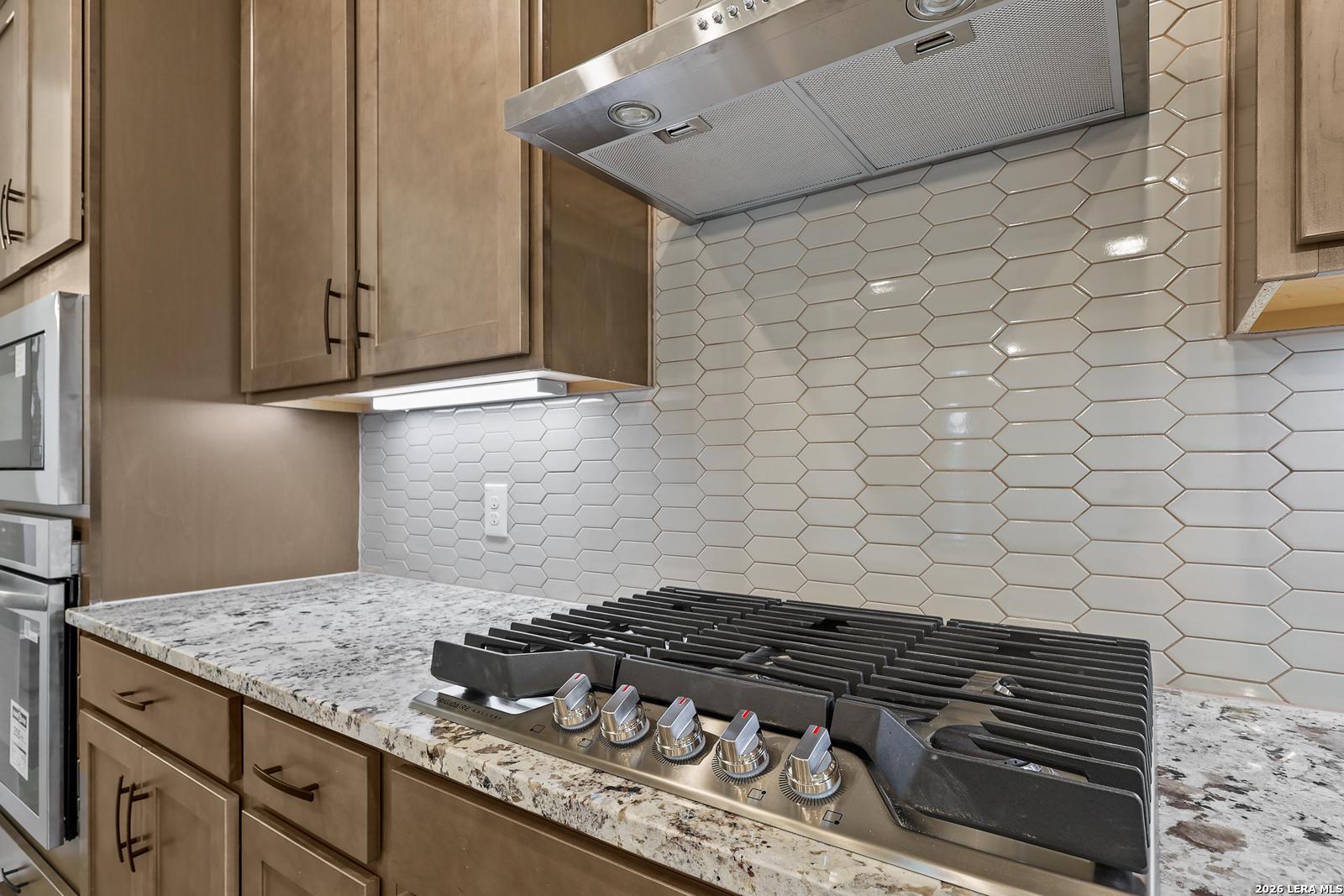 Gas cooktop with stainless hood, hexagon tile backsplash, and granite countertops in Davidson Homes Glenwood E kitchen, San Antonio