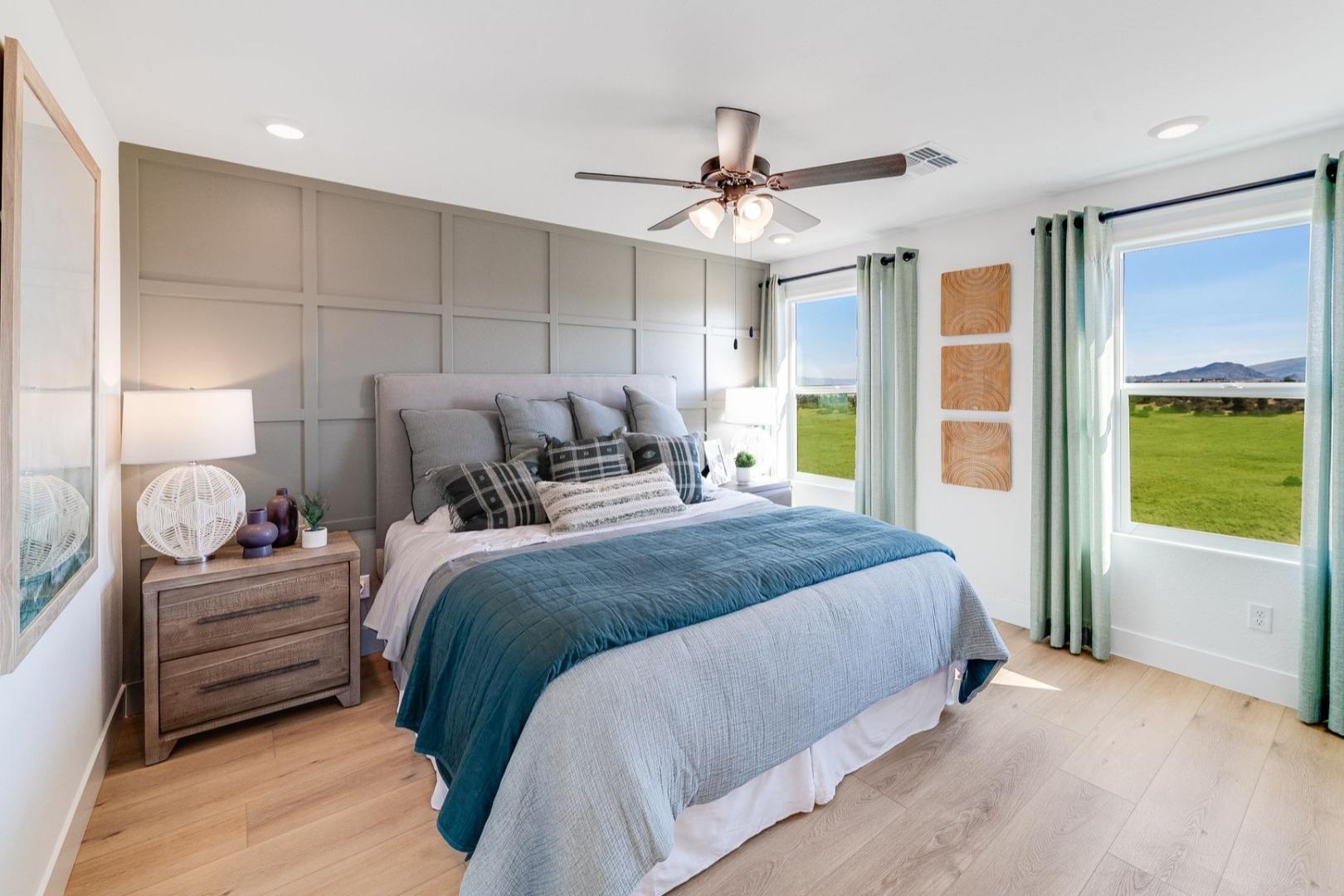 Spacious master bedroom in The Wilmington with king bed, gray accent wall, ceiling fan, and panoramic green field views through large windows