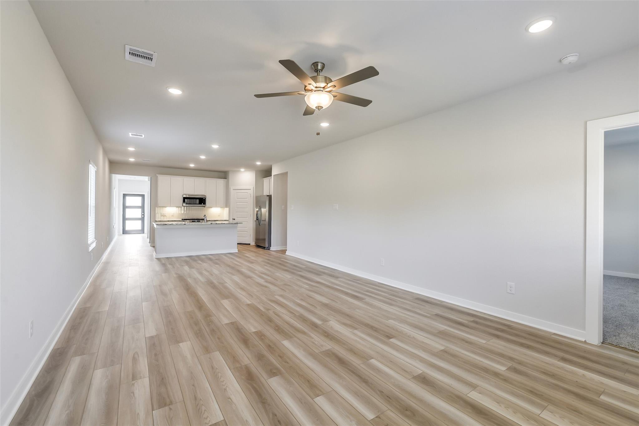 Spacious open hallway with light wood laminate floors, ceiling fan, and adjacent white kitchen cabinets in The San Marcos E, Conroe, Texas