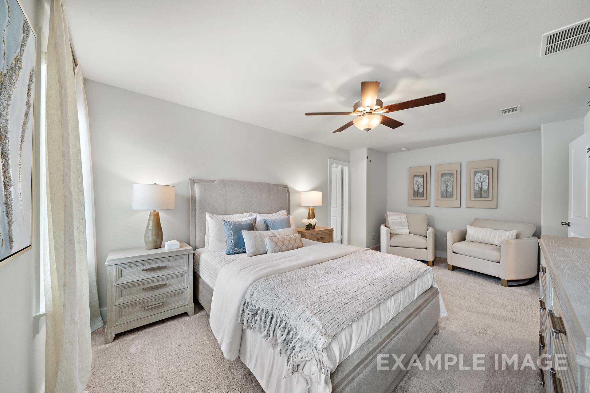 Spacious master bedroom in The Lily A featuring gray upholstered king bed, neutral armchairs, ceiling fan, and sheer curtains