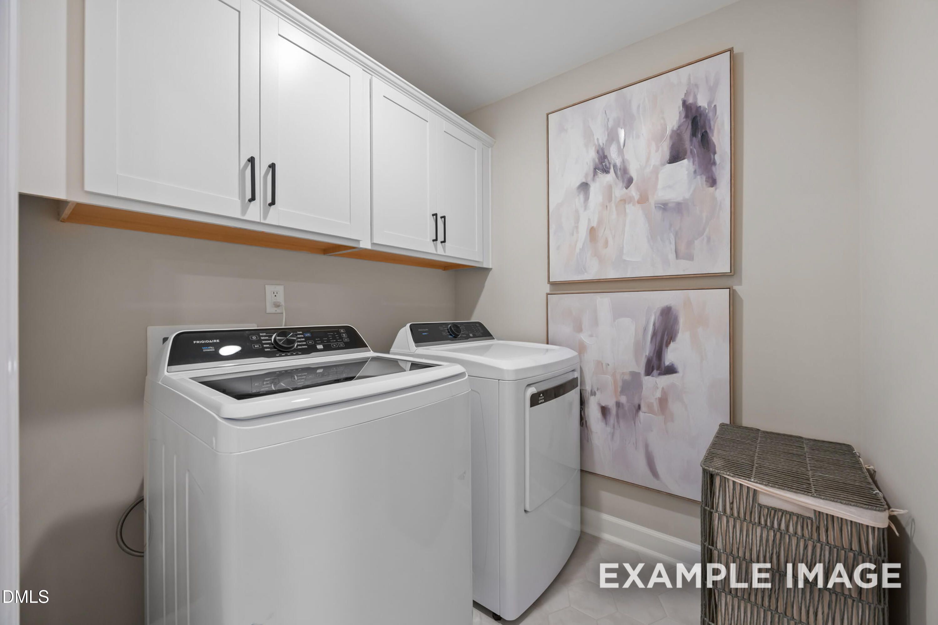 Modern laundry room with white Frigidaire washer dryer, overhead cabinets, abstract art, and wicker basket in The Mitchell home, Knightdale, NC