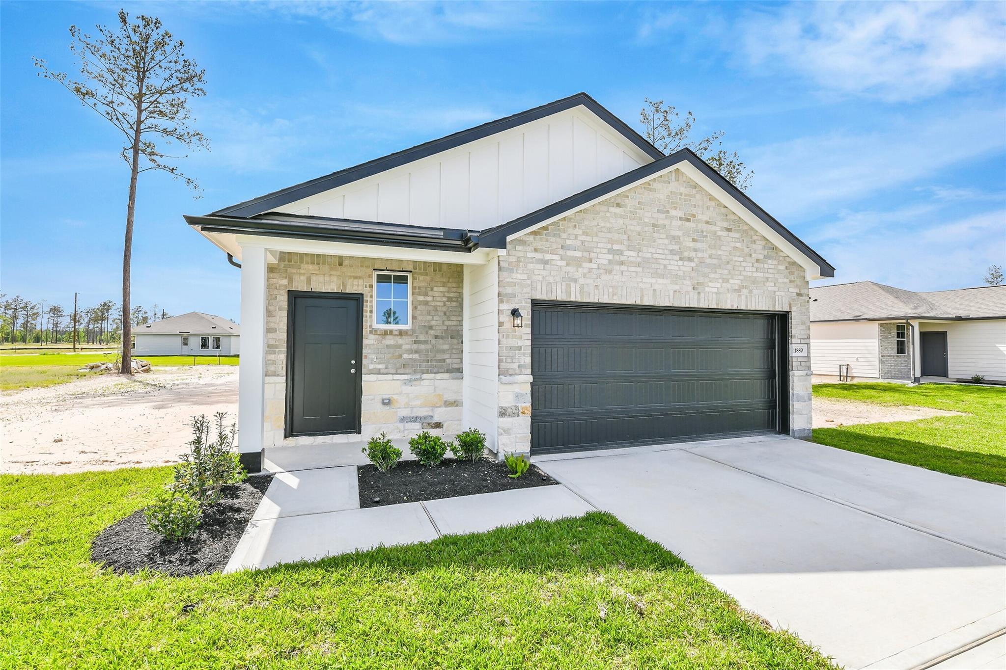 Modern beige brick single-story home with black 2-car garage, shrubs, driveway, and lush lawn in Spring Branch Crossing, Conroe, Texas - The Frio G by Davidson Homes